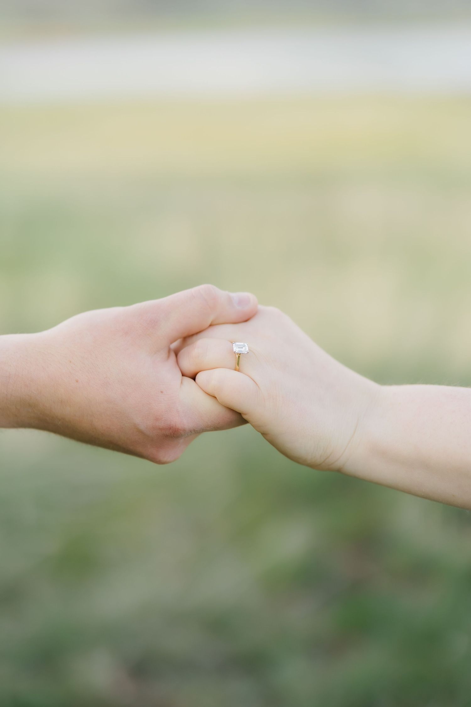 Close-up of engaged couple holding hands showing a diamond engagement ring outdoors.