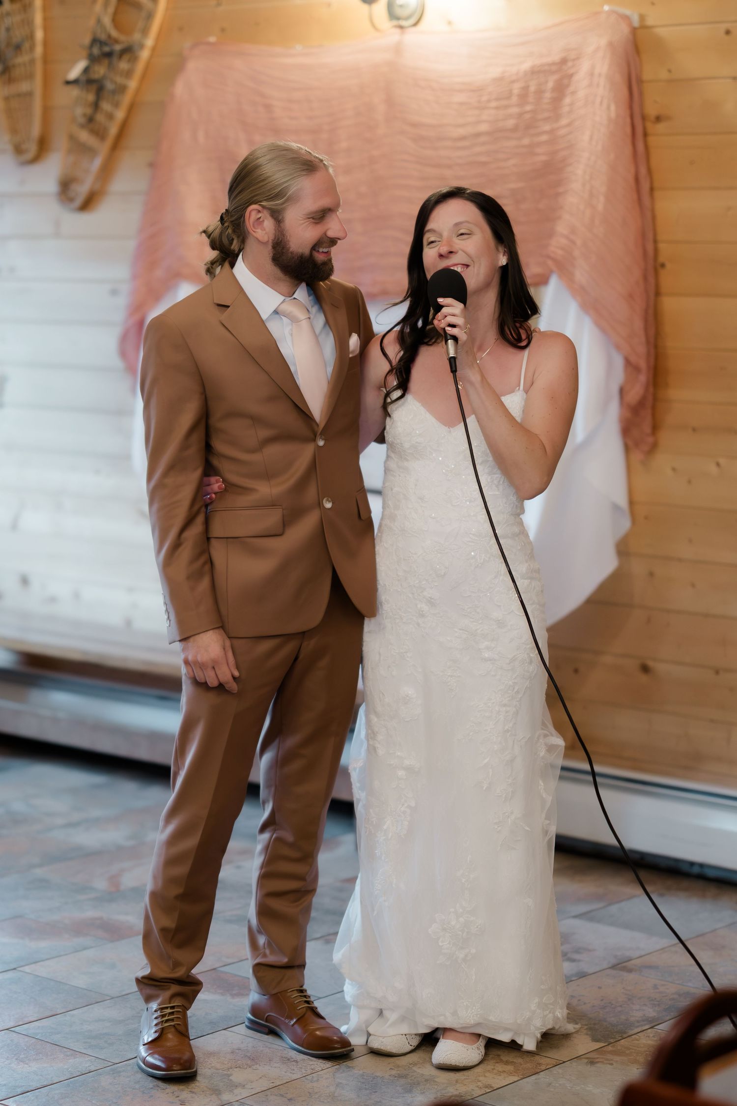 Bride in white lace gown speaks into microphone beside groom in tan suit during wedding reception speech.