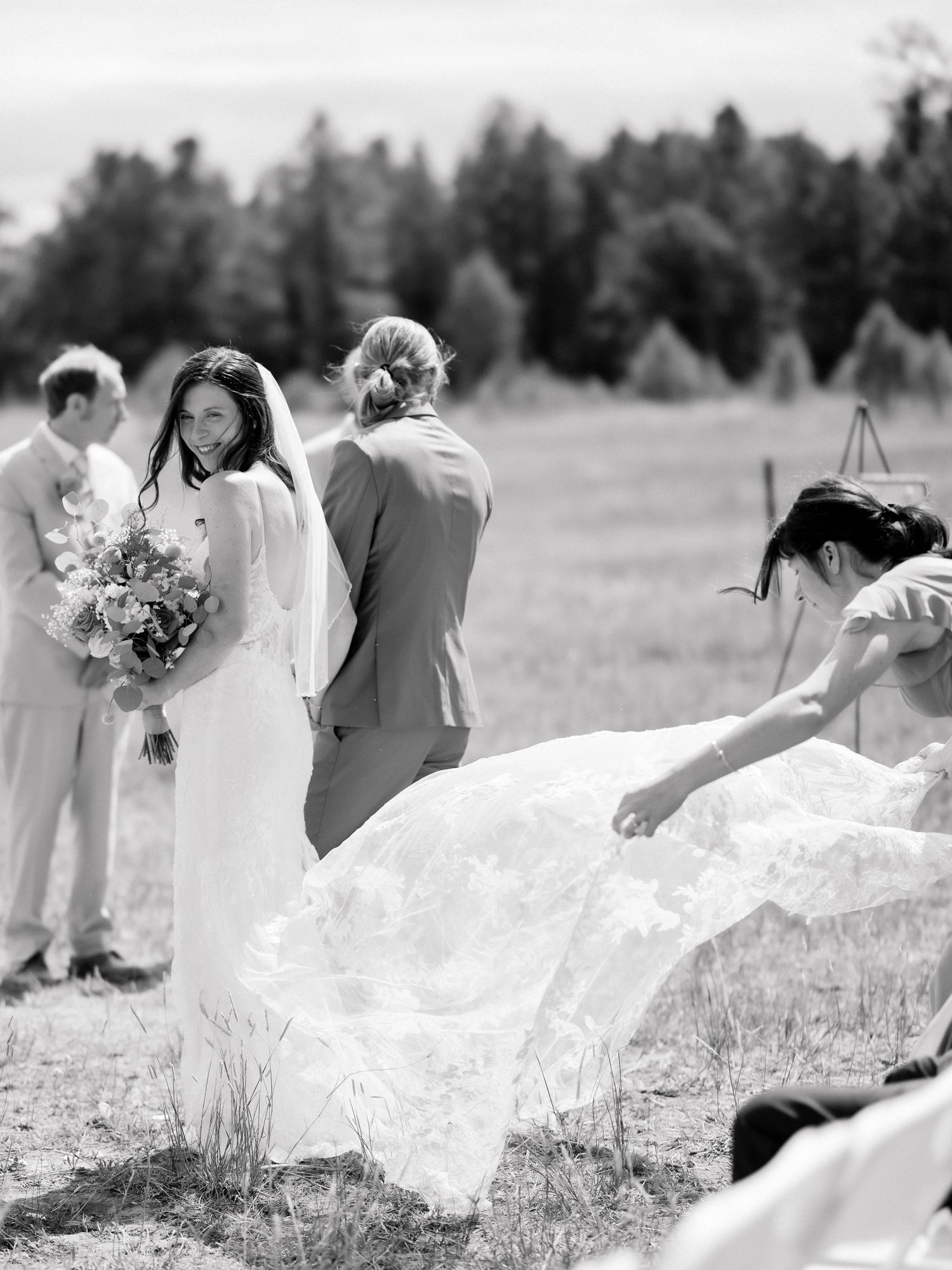 Black and white photo of a bride holding bouquet as attendant arranges her lace gown train at outdoor wedding ceremony.