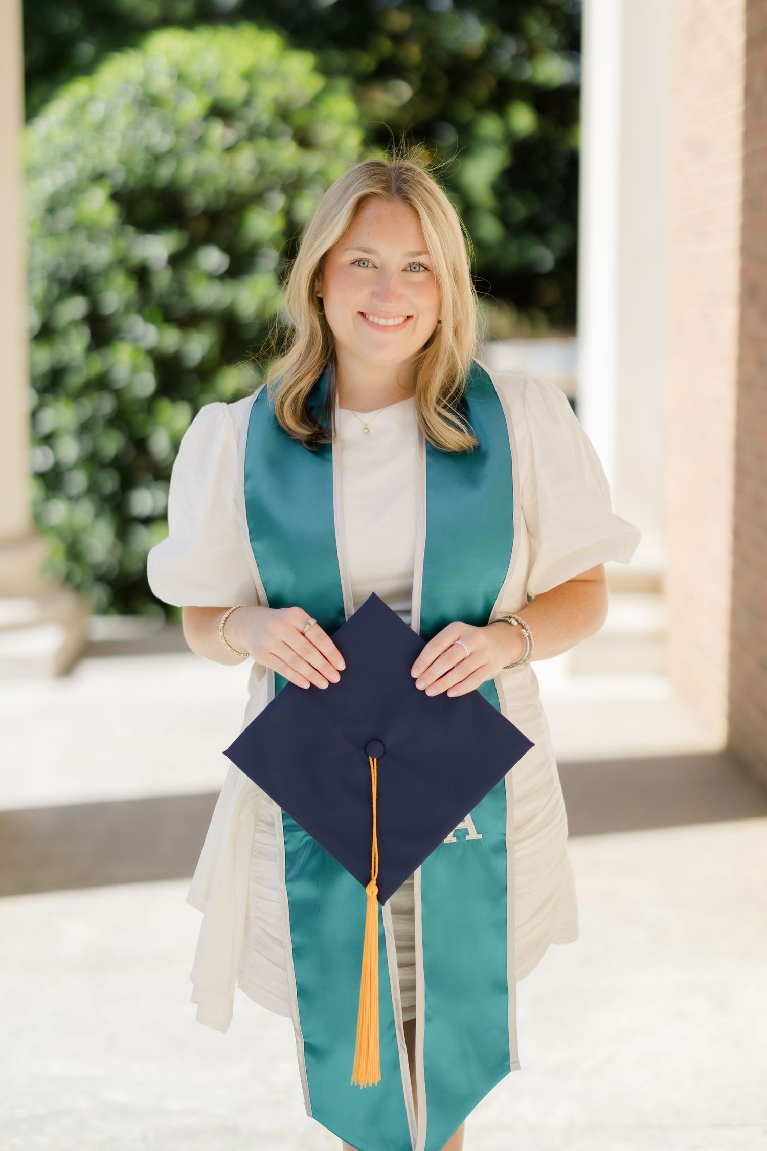 Smiling blonde woman in teal graduation stole holding navy mortarboard cap outdoors at graduation ceremony.