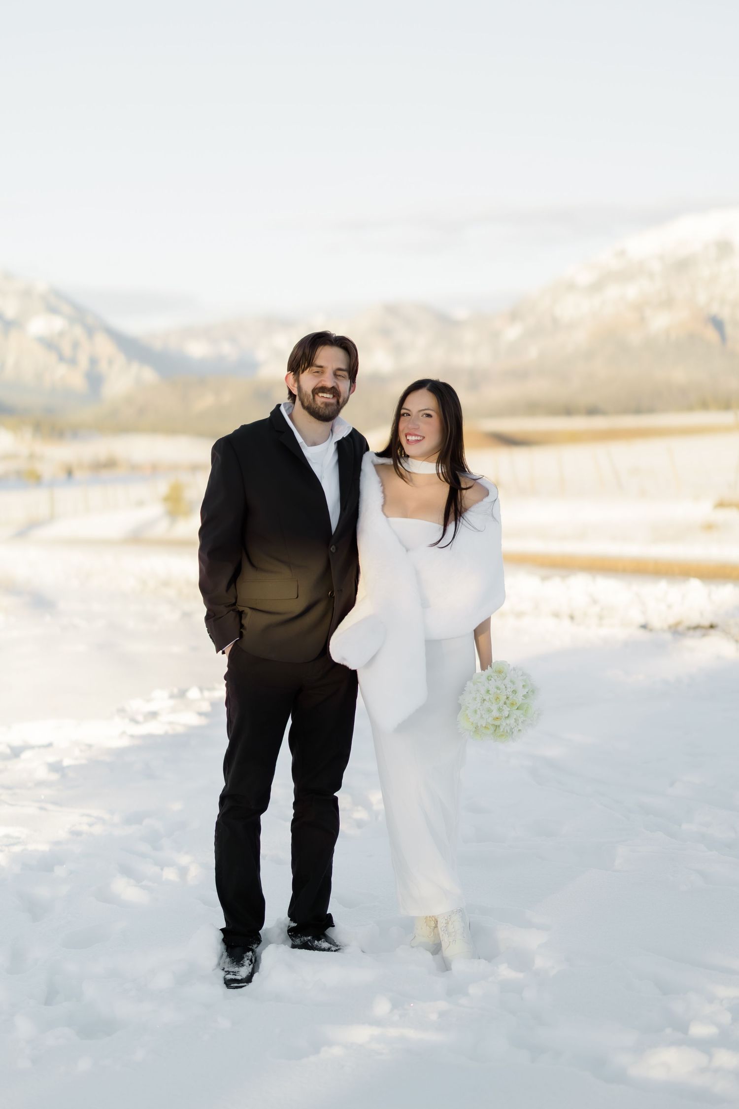Smiling wedding couple poses in snow-covered mountain valley, bride in white dress with fur wrap.