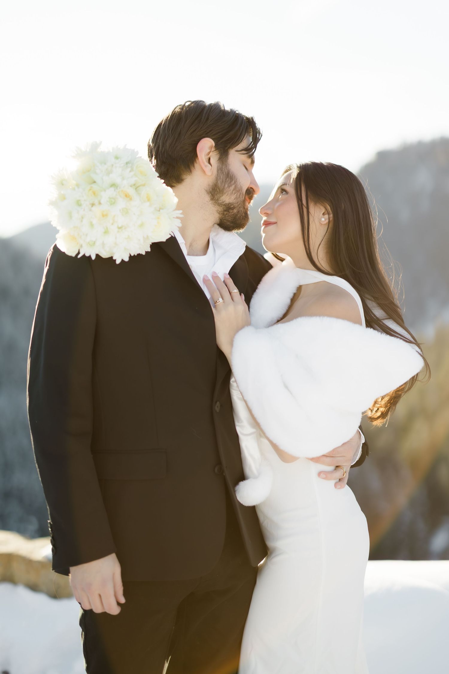 Bride and groom share a romantic moment outdoors in winter, she in white dress and fur stole, he in black suit.