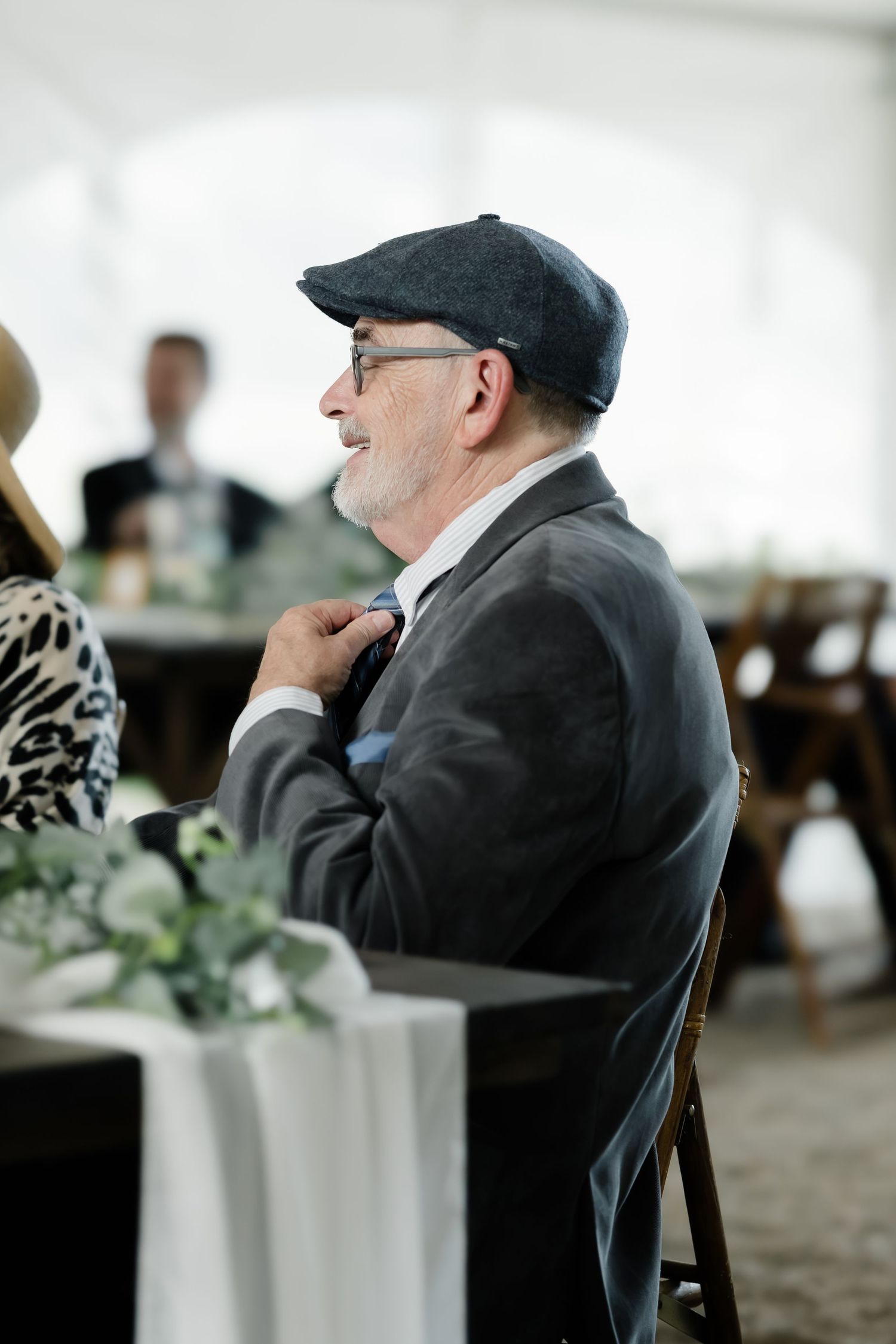 A smiling elderly man in a flat cap and gray suit adjusts his bow tie at a wedding reception.