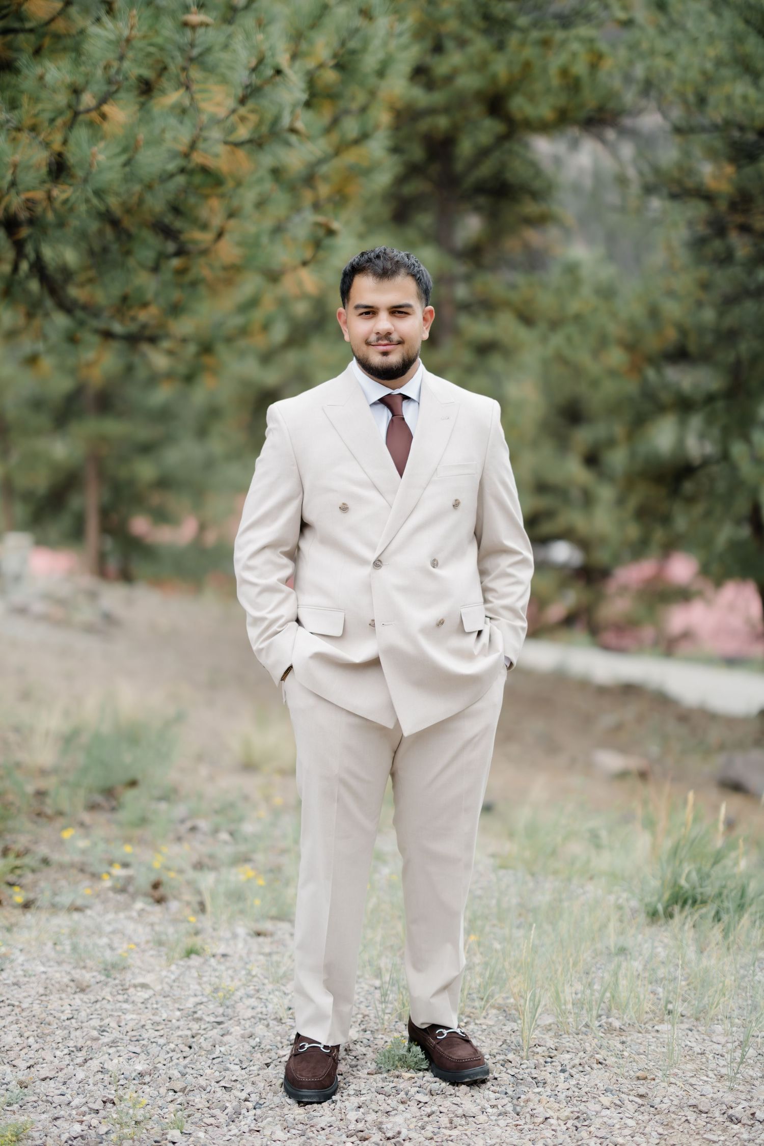 A groom in a beige double-breasted suit with a burgundy tie stands outdoors among pine trees.