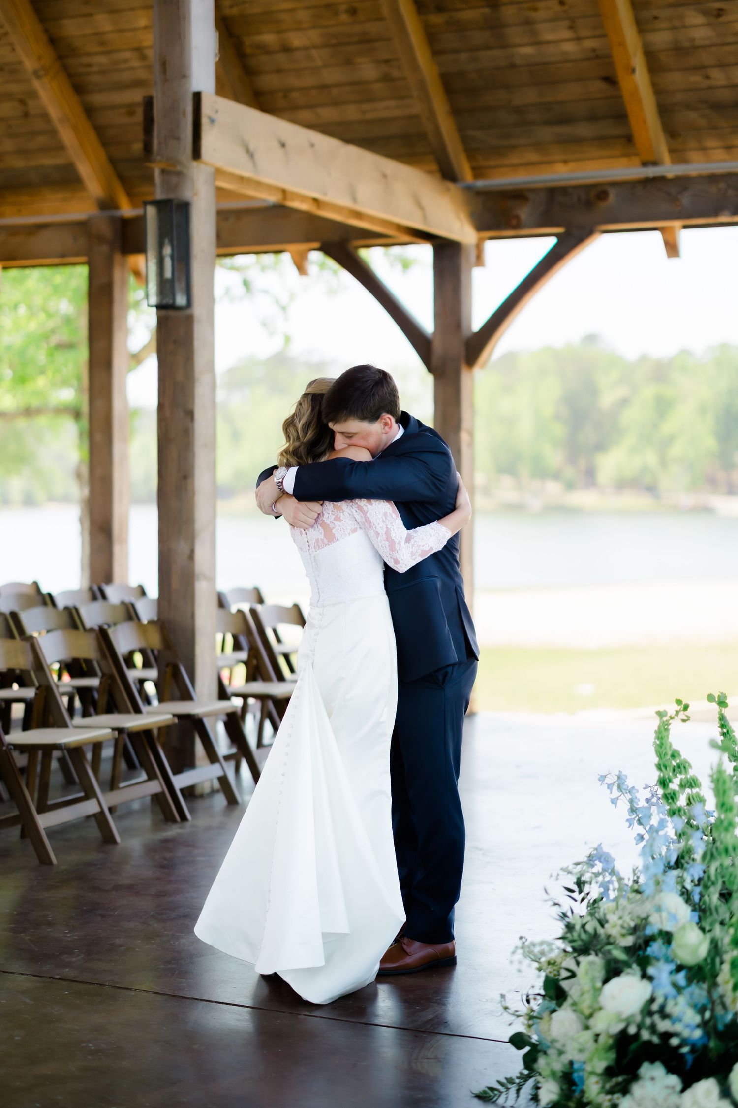 Bride and groom share a heartfelt embrace at an outdoor rustic wedding venue with floral arrangements.