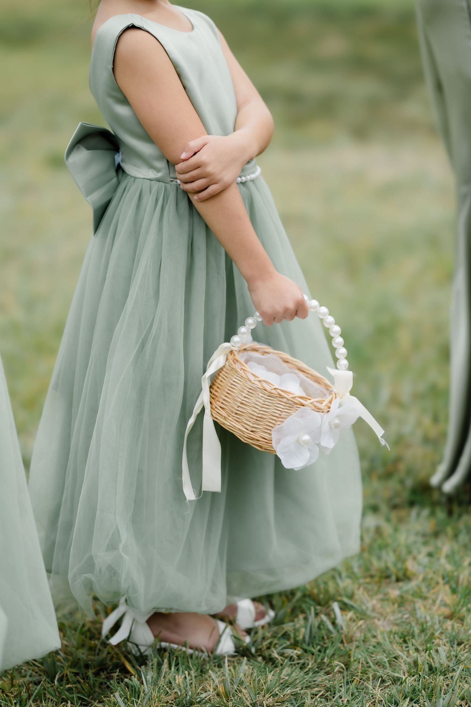 A flower girl in a sage green tulle dress holds a wicker basket with pearl handle and white ribbon at an outdoor wedding.