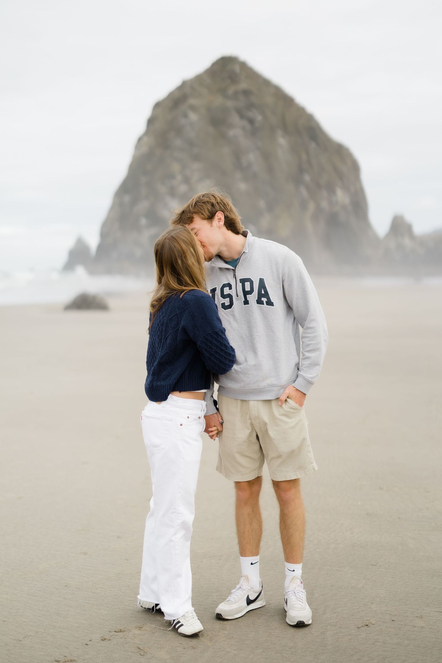 Couple sharing a kiss on a misty beach with Haystack Rock in the background at Cannon Beach, Oregon.