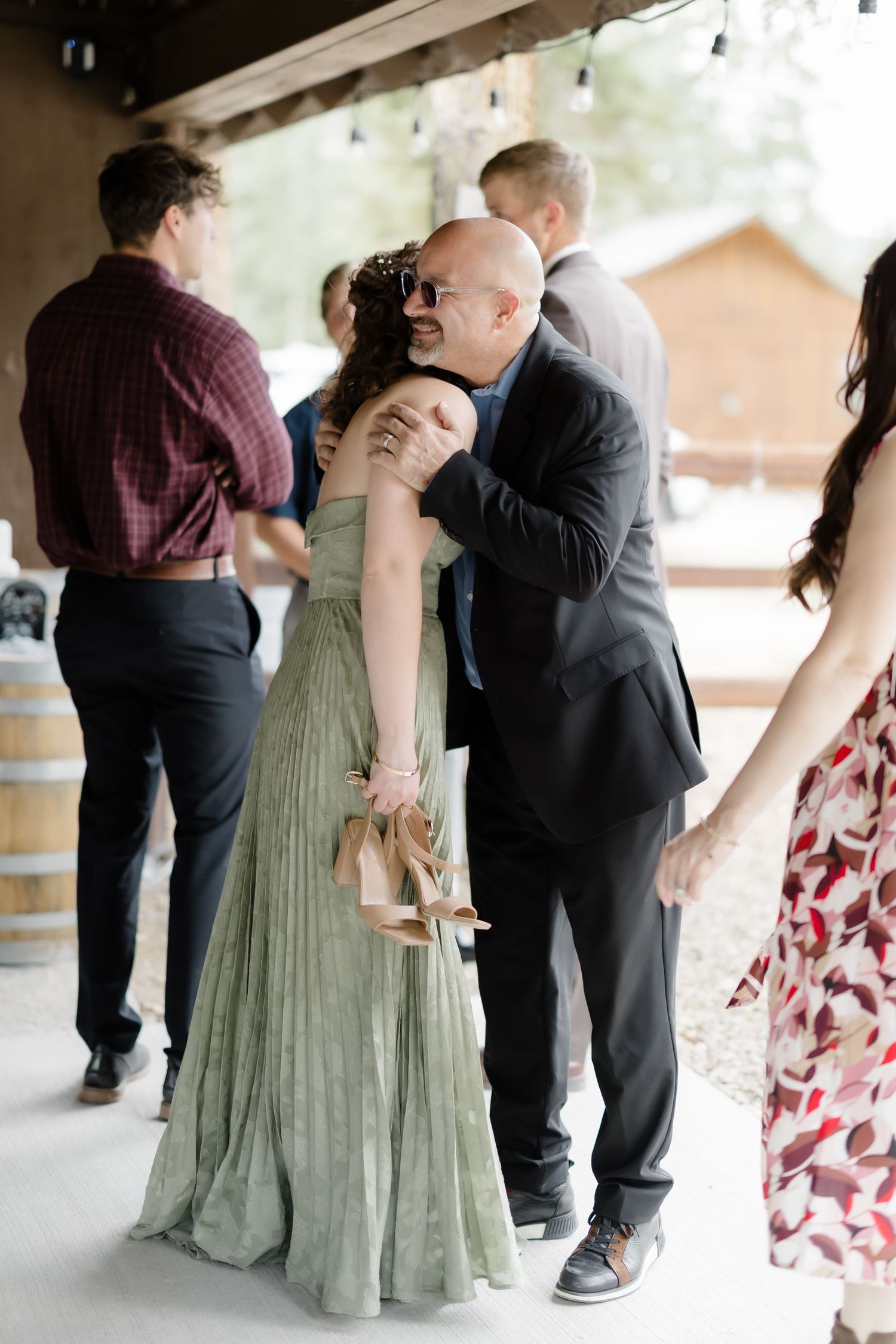 A bald man in a black suit warmly embraces a woman in a sage green pleated gown at an outdoor wedding reception.