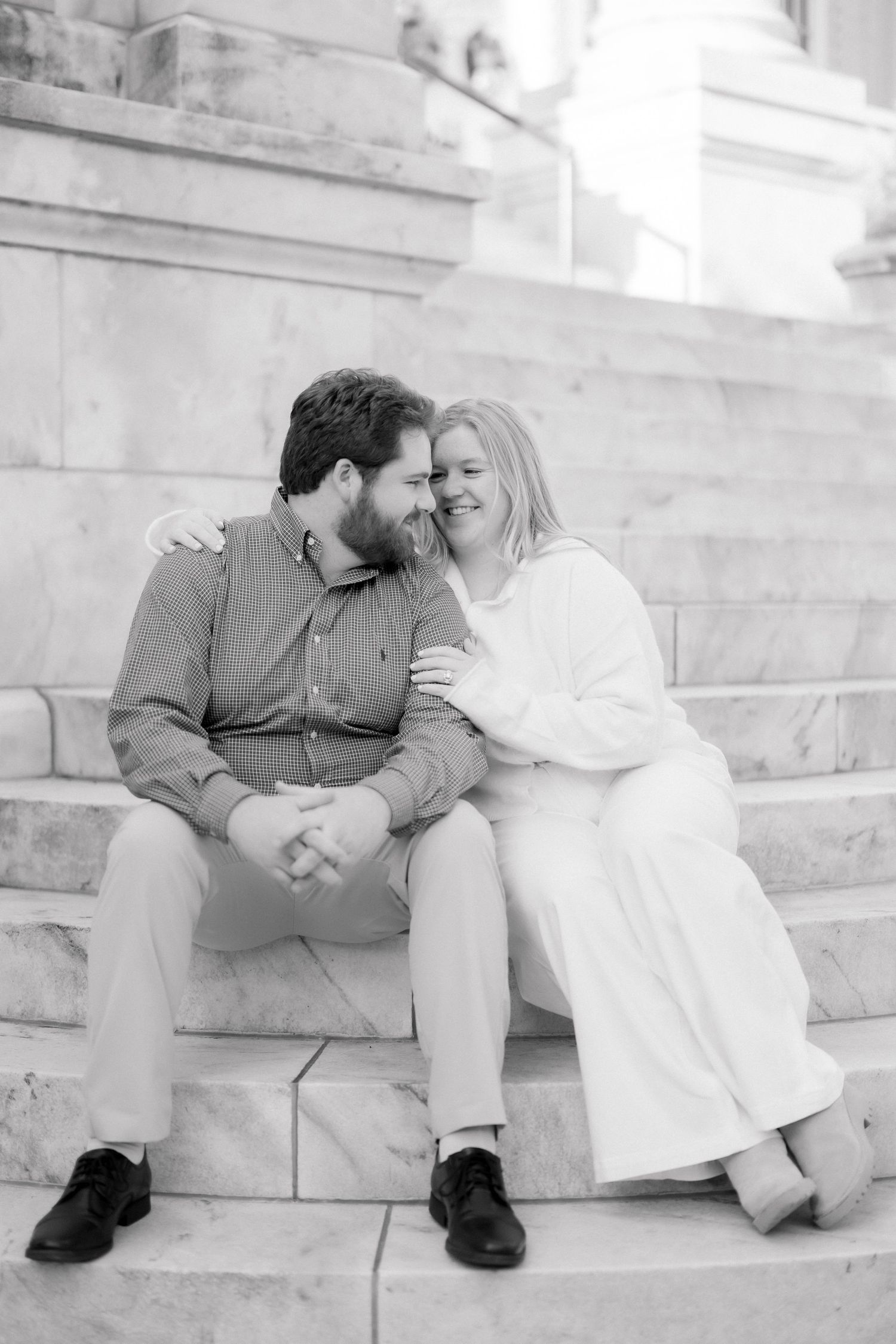 Black and white photo of a couple sitting on marble steps, smiling and embracing during an engagement session.