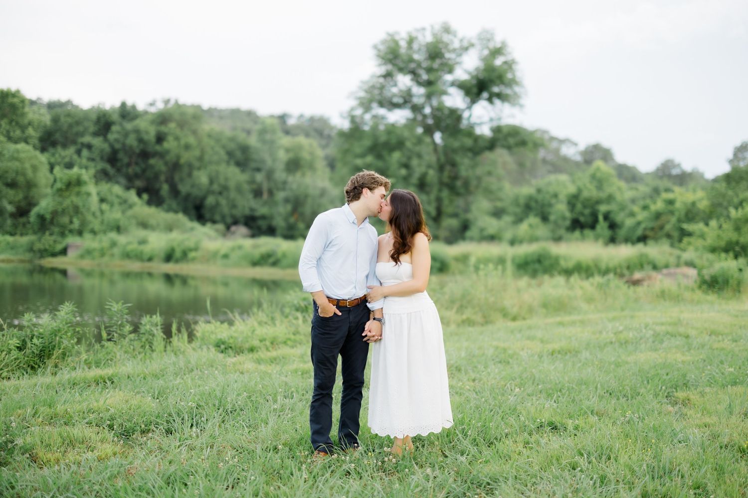 A couple shares a romantic kiss in a lush green meadow by a serene river during their outdoor engagement session.