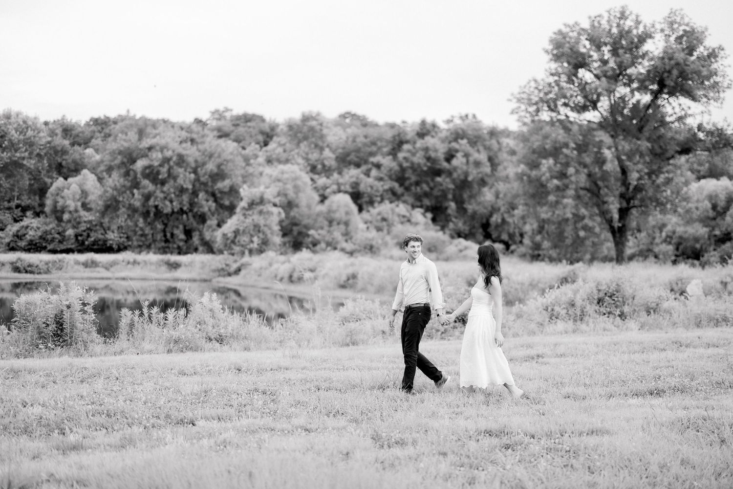 Black and white photo of a couple holding hands walking through an open field near a serene pond.