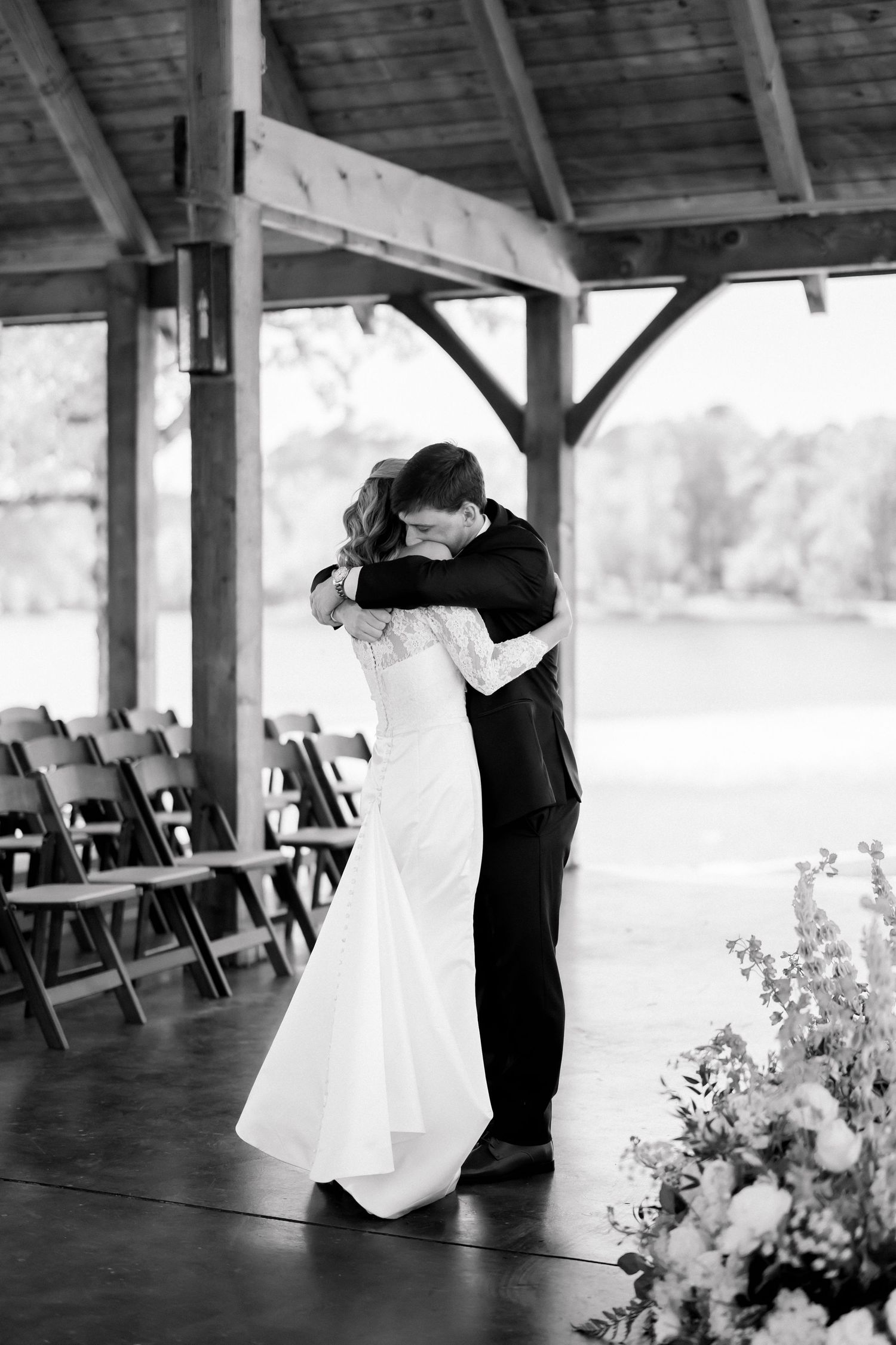 Black and white photo of bride and groom sharing an emotional first dance embrace under a rustic wooden pavilion.