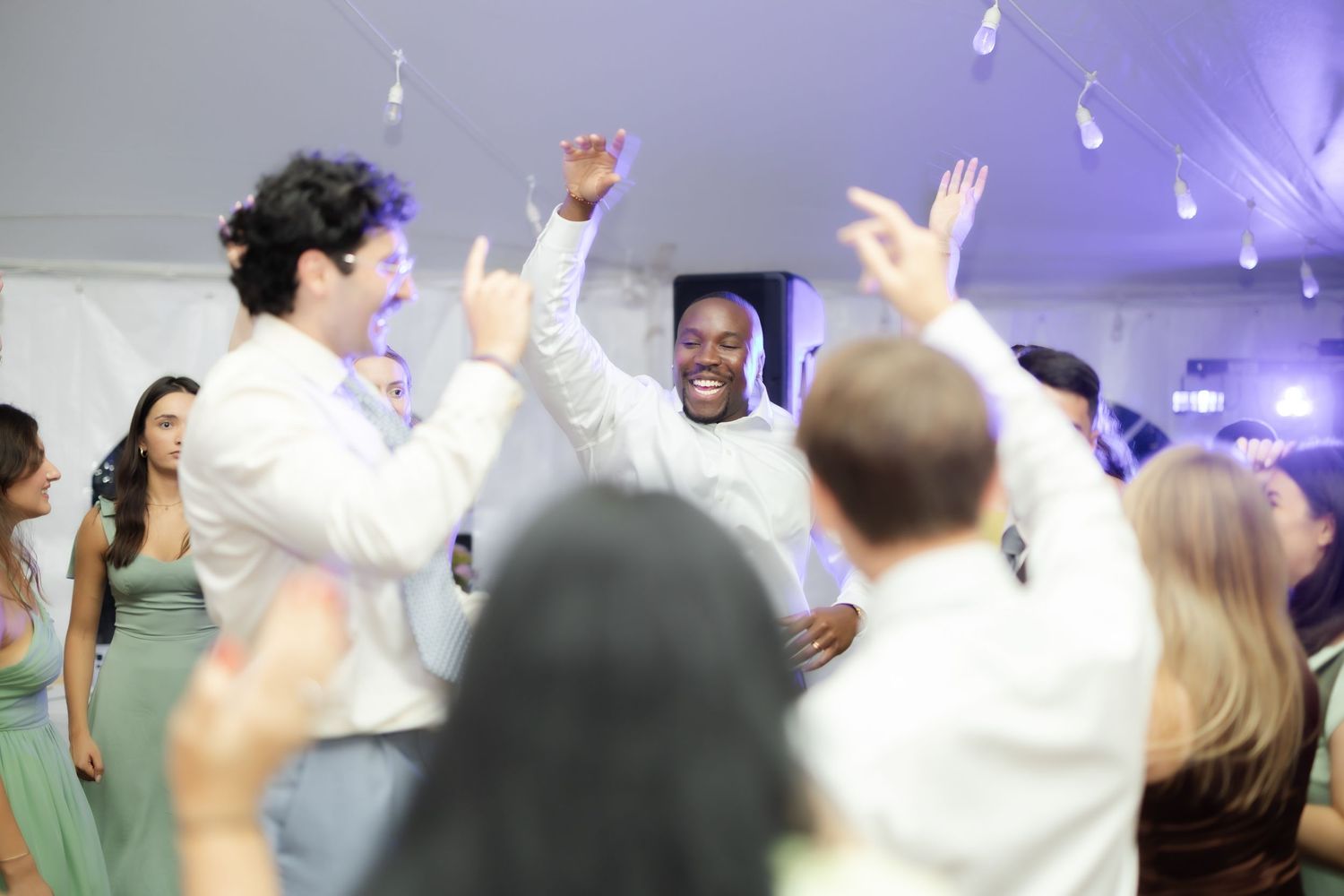 Guests dancing joyfully at a wedding reception under a white tent, with hands raised in celebration.