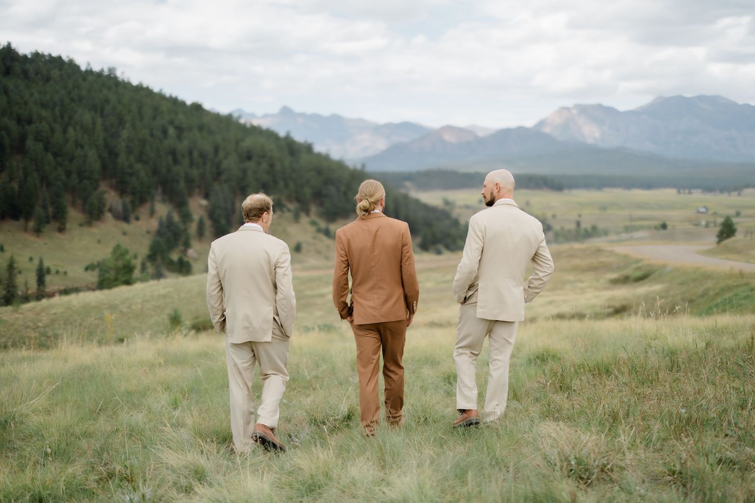 Three groomsmen in tan and brown suits walk through a scenic mountain meadow with evergreen trees and peaks in the background.