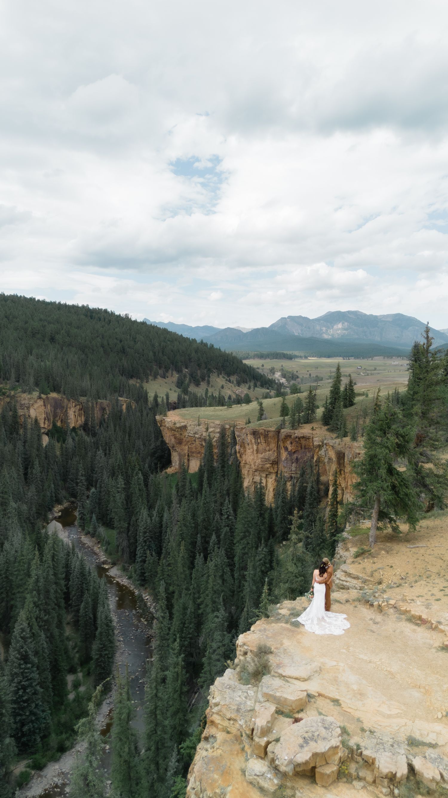 Bride in white gown stands on rocky cliff edge overlooking a forested canyon and mountain valley under cloudy skies.