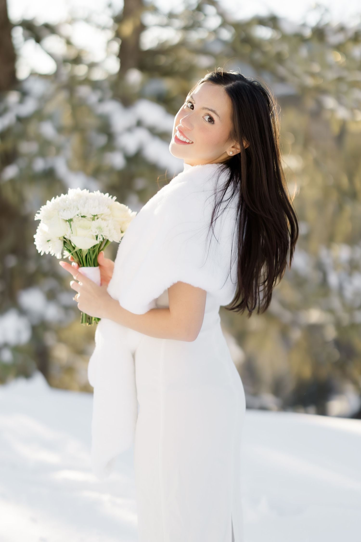 Smiling bride in white dress and fur wrap holds white flower bouquet in snowy winter setting.
