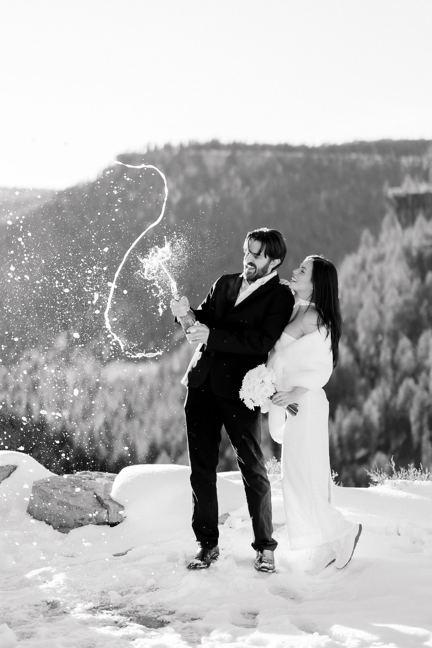 Black and white photo of newlyweds popping champagne bottle in snowy mountain setting.