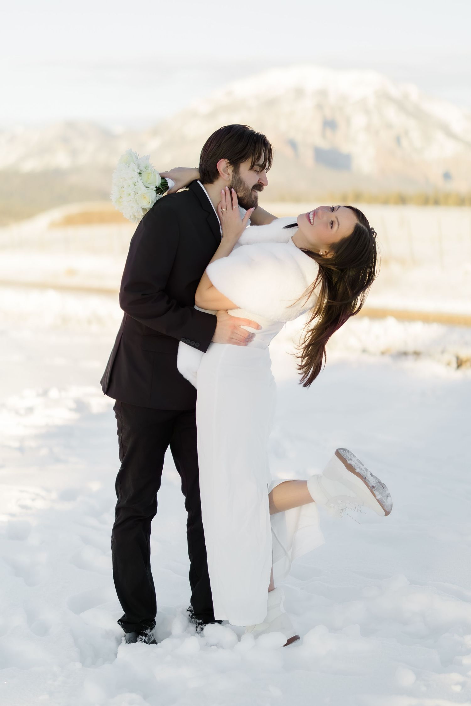 Groom lifts smiling bride in white dress and fur wrap in snowy mountain setting during winter wedding.