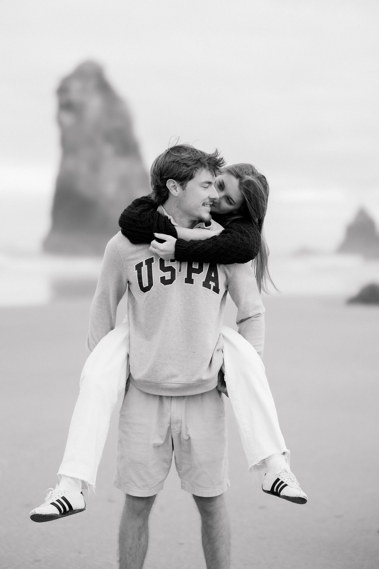 Black and white photo of a couple on a beach, man carrying woman piggyback, wearing USPA sweatshirt.