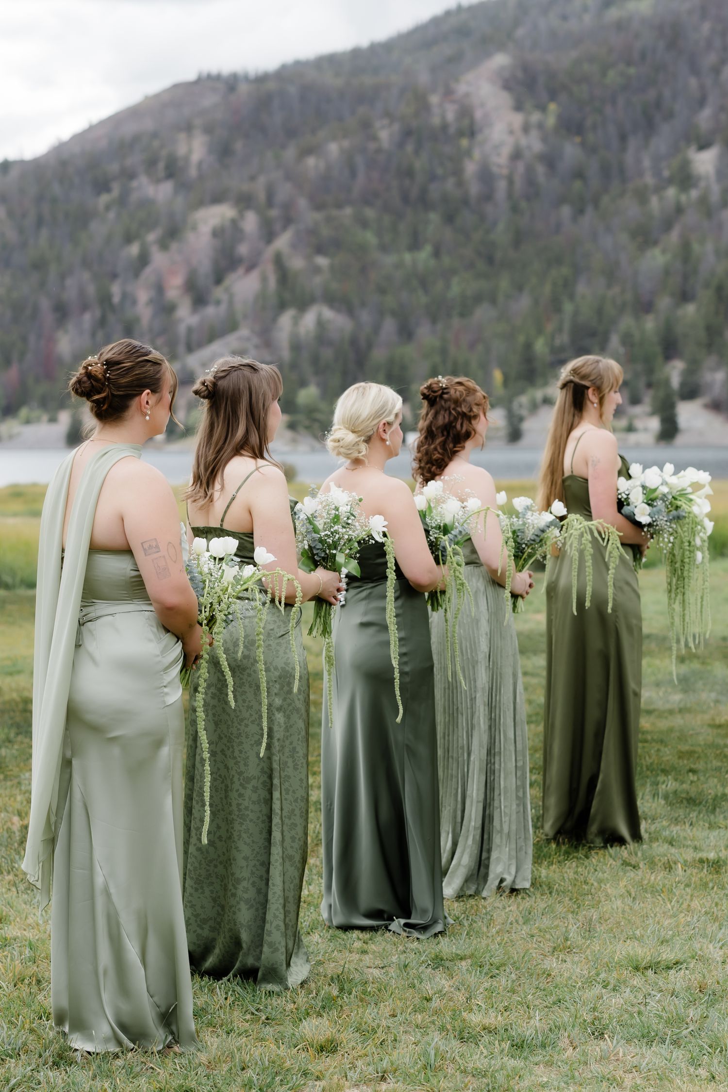 Four bridesmaids in sage green gowns holding white bouquets, standing before a mountain lake backdrop.