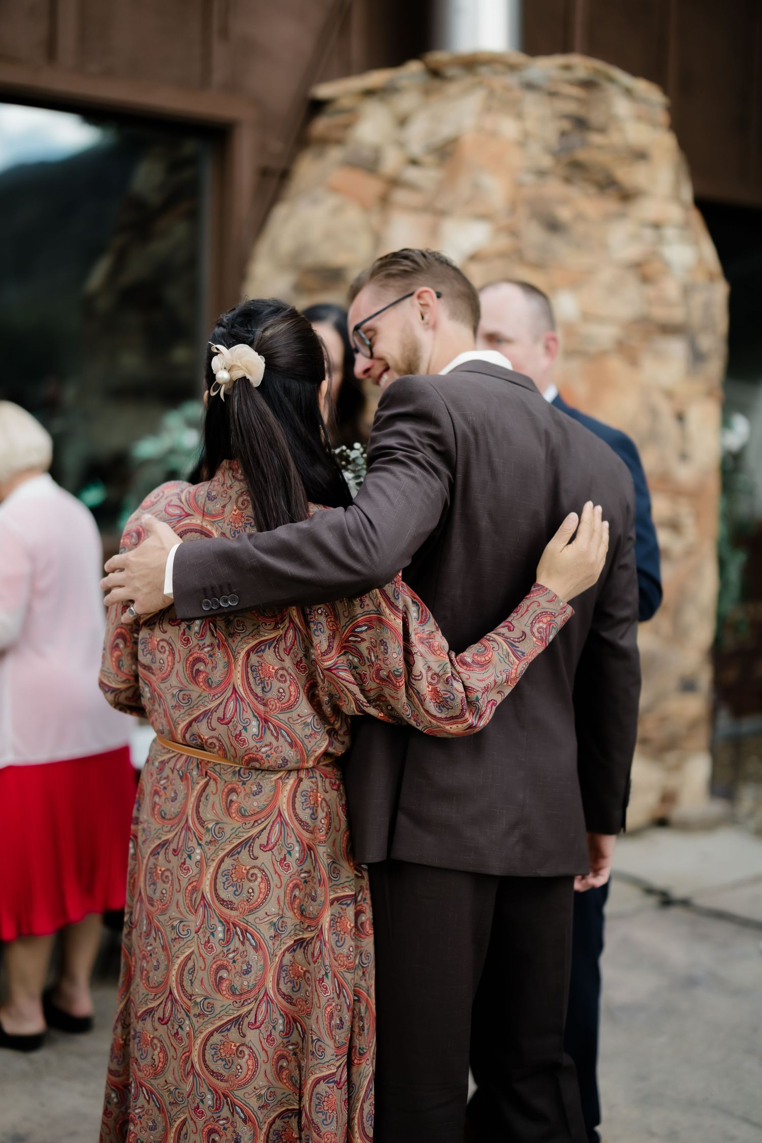 A groom in a brown suit embraces a woman in a paisley dress at an outdoor wedding venue.