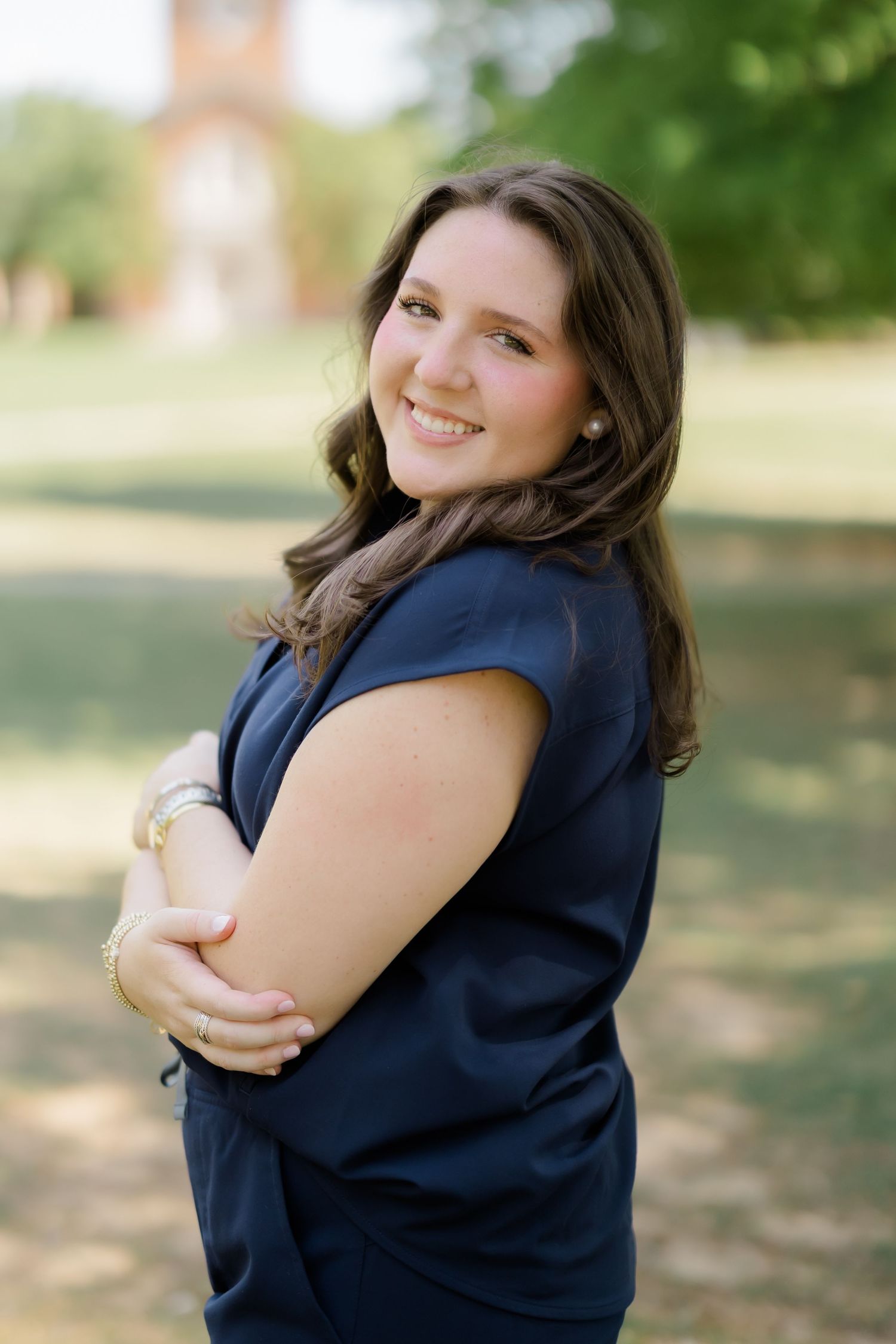 A smiling young woman with brown hair in a navy dress poses outdoors on a sunny day in a park setting.