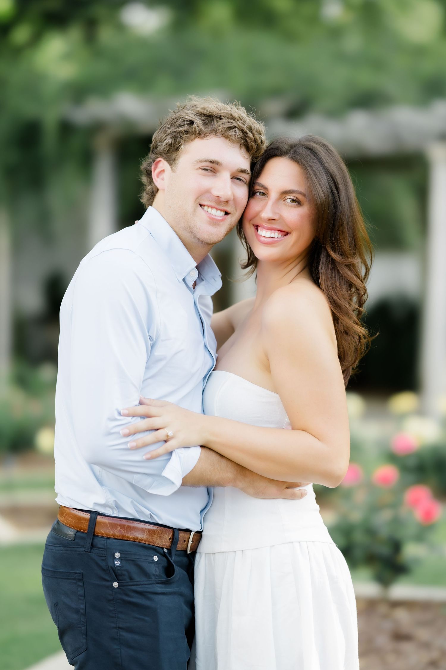 Smiling couple embracing outdoors in a garden, woman in white dress and man in light blue shirt and navy pants.