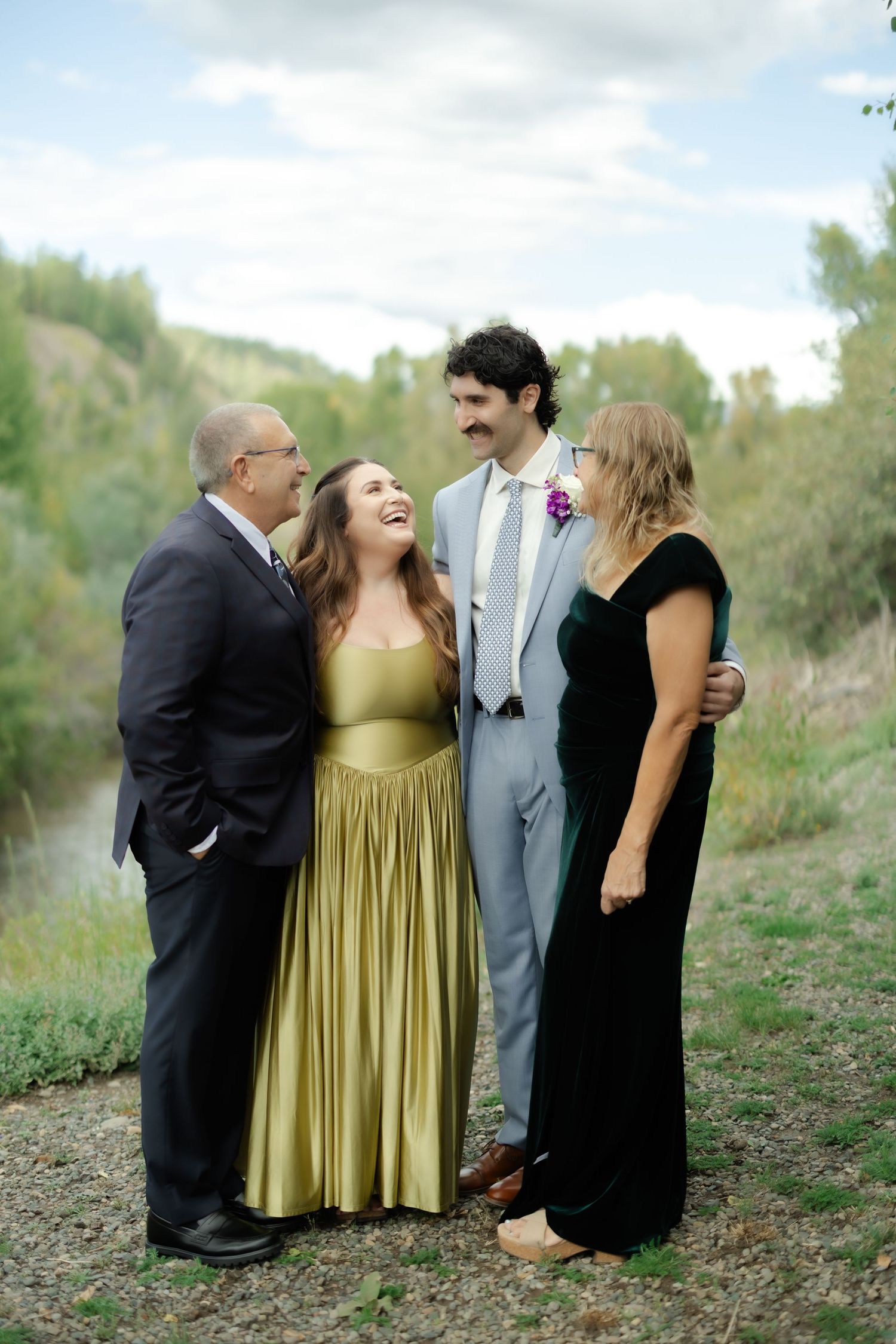 Two couples pose outdoors at a wedding, smiling together near a river with lush greenery in the background.