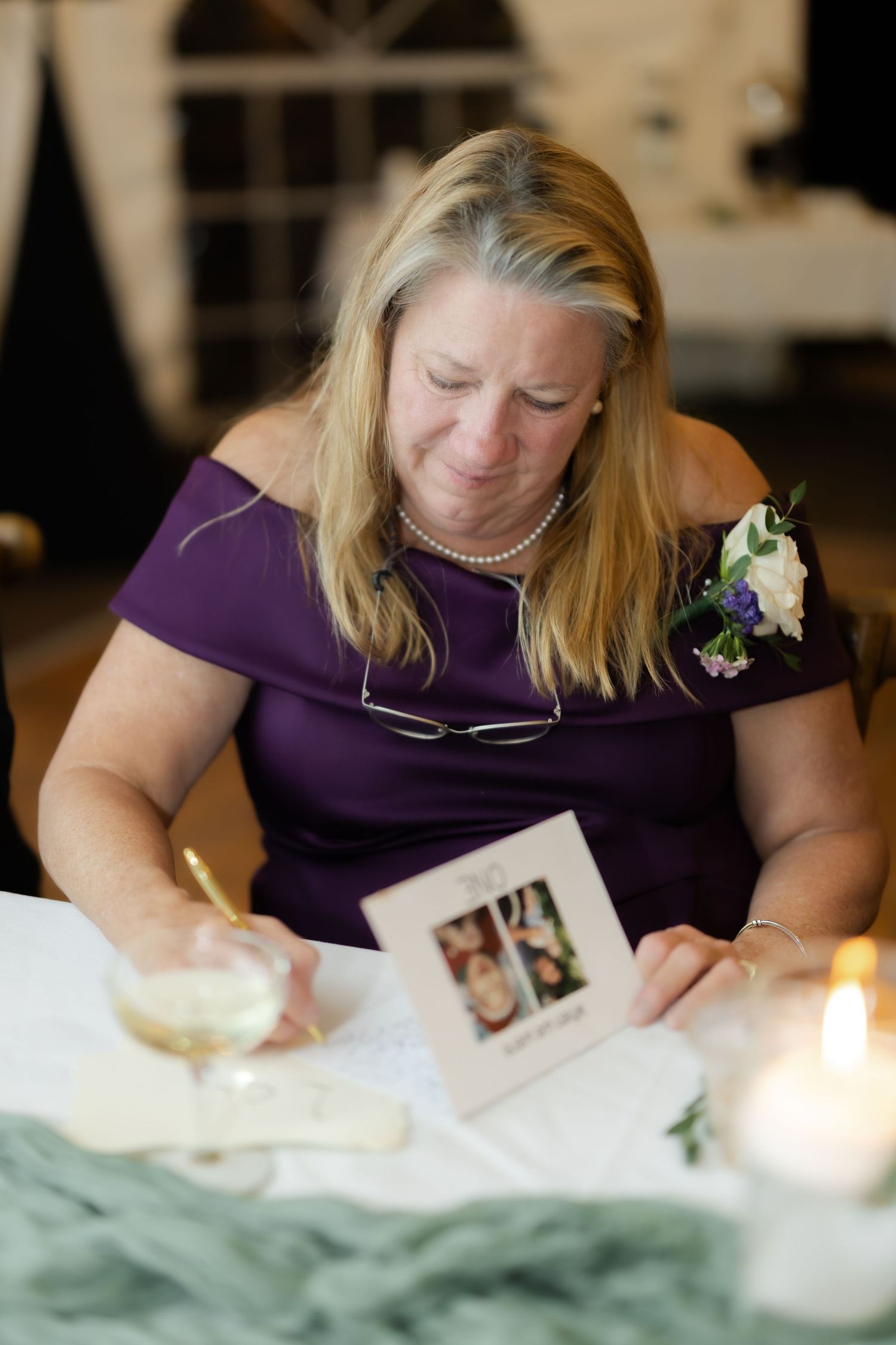 Emotional woman in purple dress with corsage writing in wedding guest book at reception table.