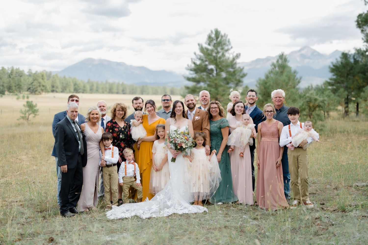 Wedding party and family pose outdoors in mountain meadow with pine trees and peaks in background.