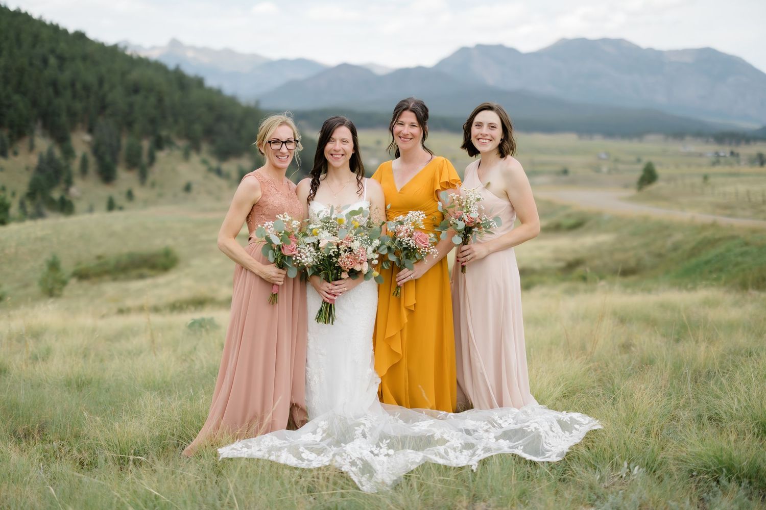 Bride and three bridesmaids in blush and yellow gowns holding bouquets in a mountain meadow.