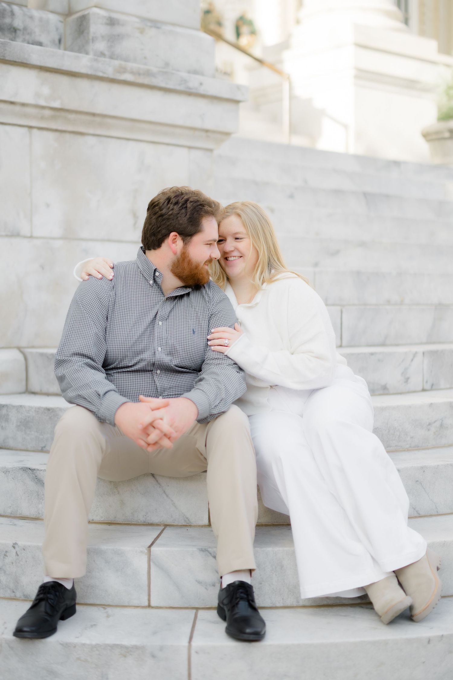 A couple sits closely on marble steps, smiling and embracing each other during an outdoor engagement photo session.