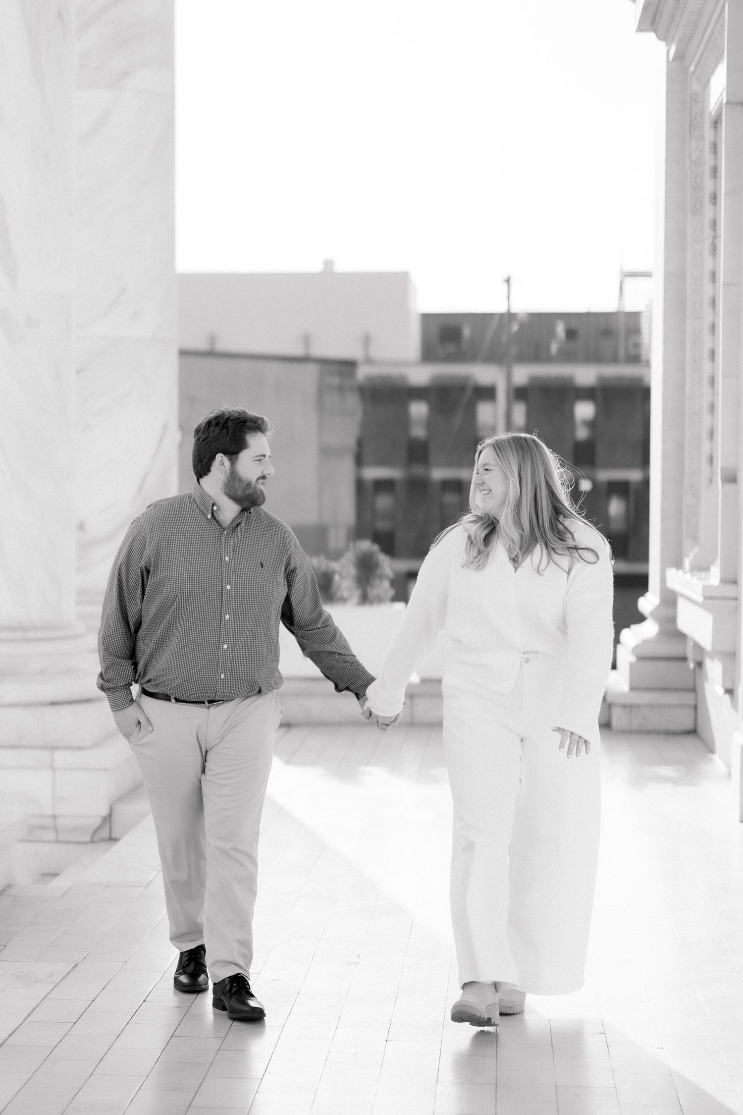 Black and white image of smiling couple holding hands and walking between large marble columns.