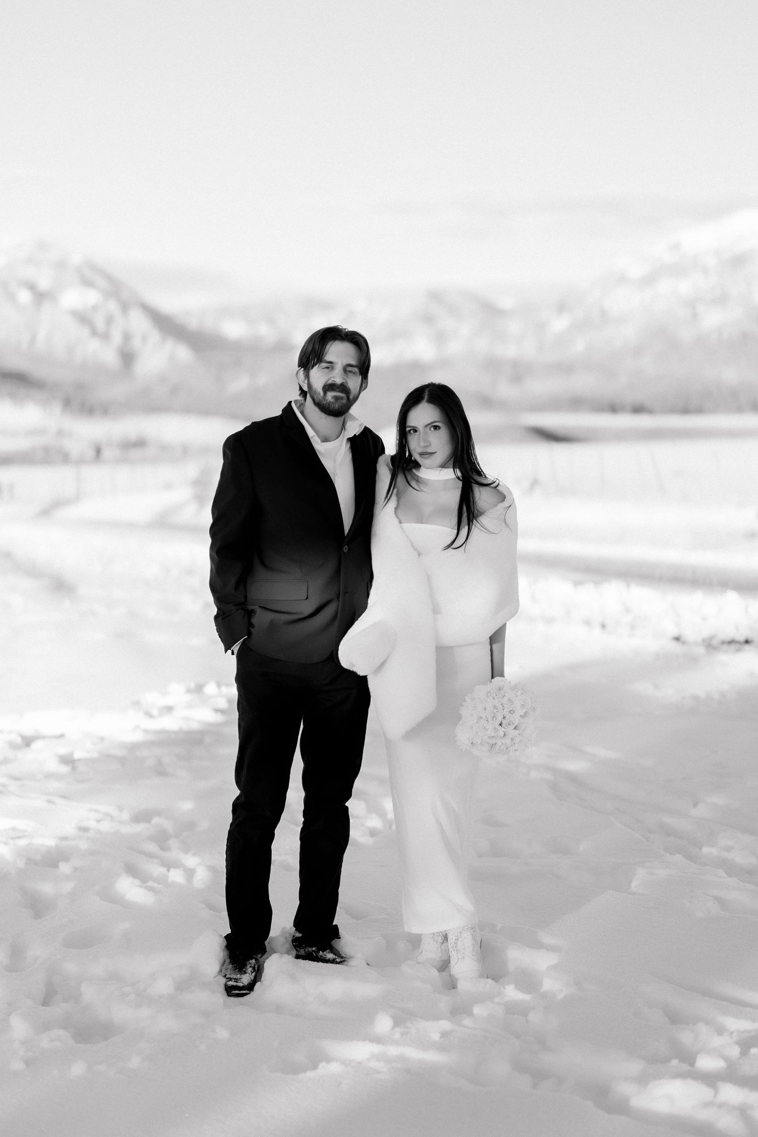 Black and white photo of bride and groom posing together in vast snowy mountain landscape.