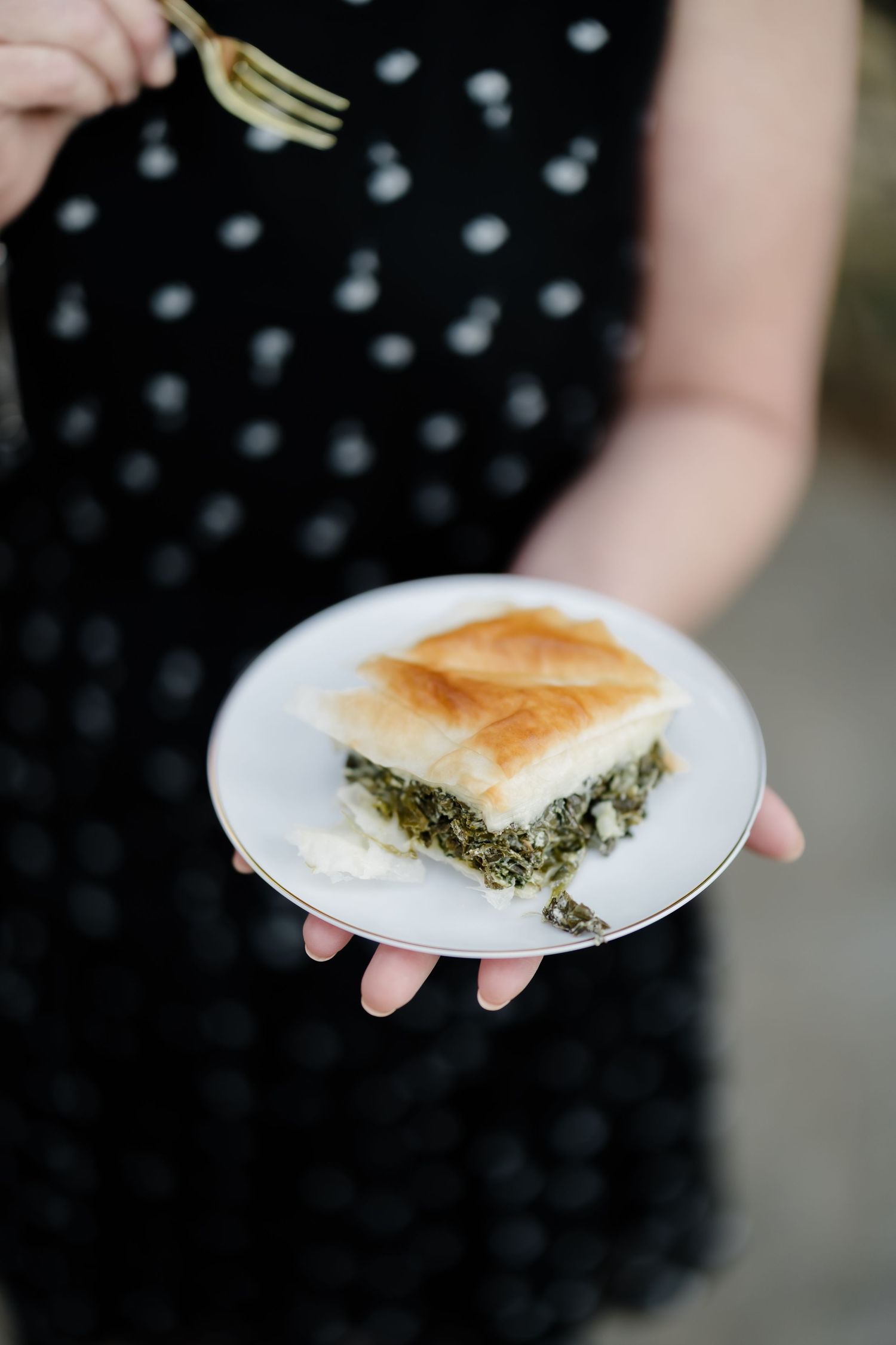 Woman in polka dot dress holding a white plate with a slice of spanakopita spinach feta phyllo pie.