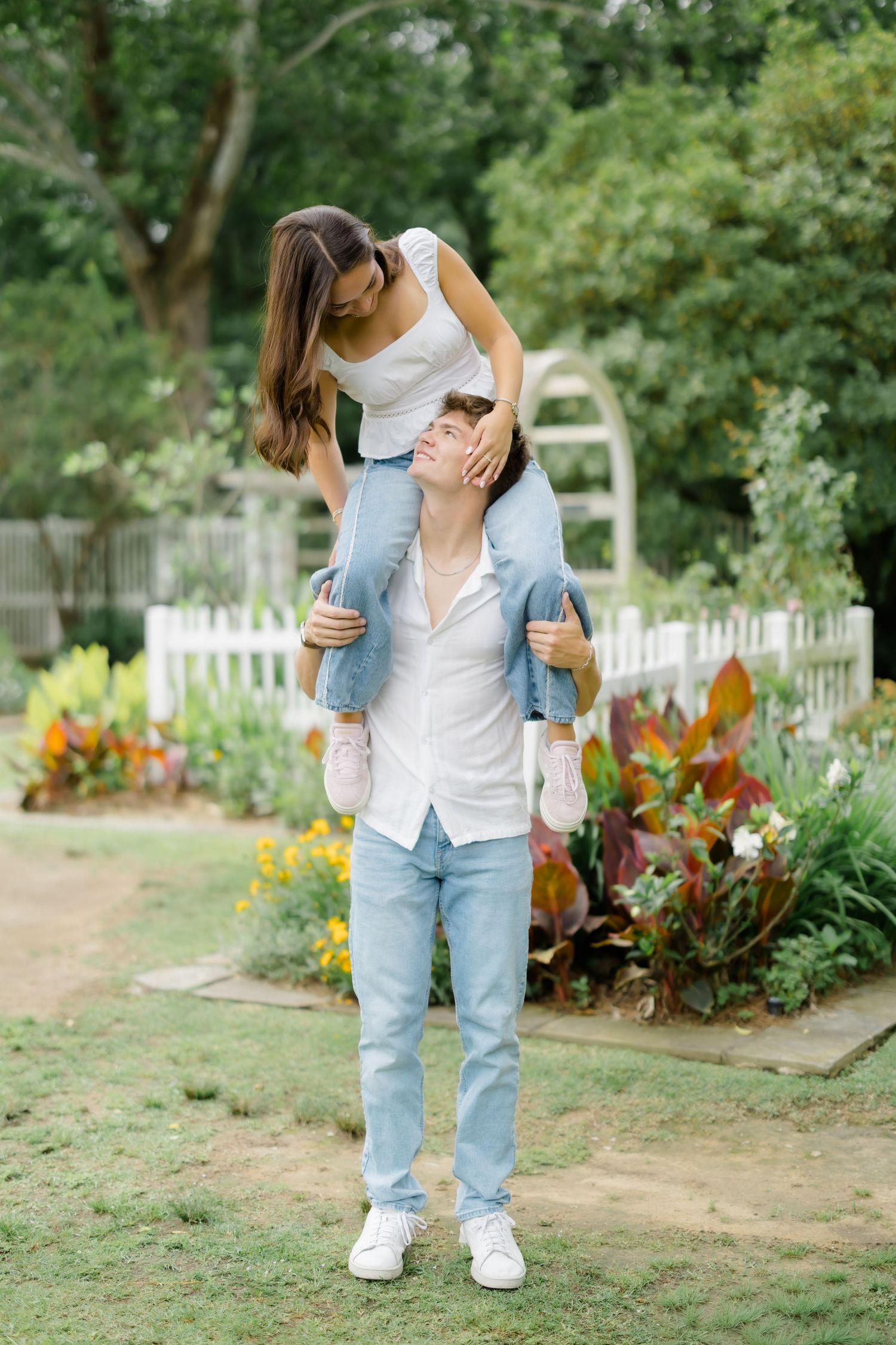 Young couple in a garden, man carrying woman on his shoulders, both smiling, surrounded by colorful flowers.