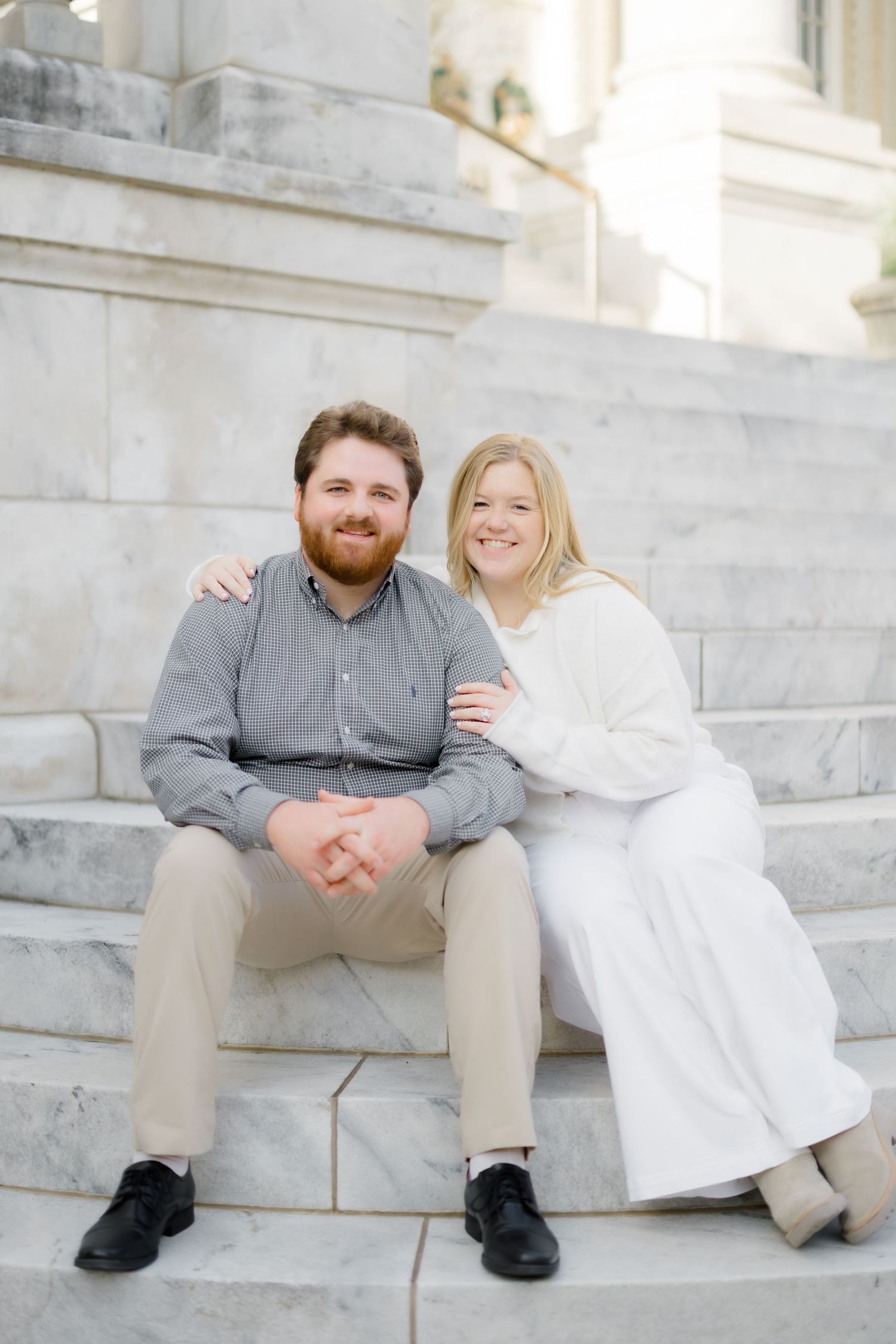 Smiling engaged couple sitting together on marble steps, woman in white dress, man in gray shirt.