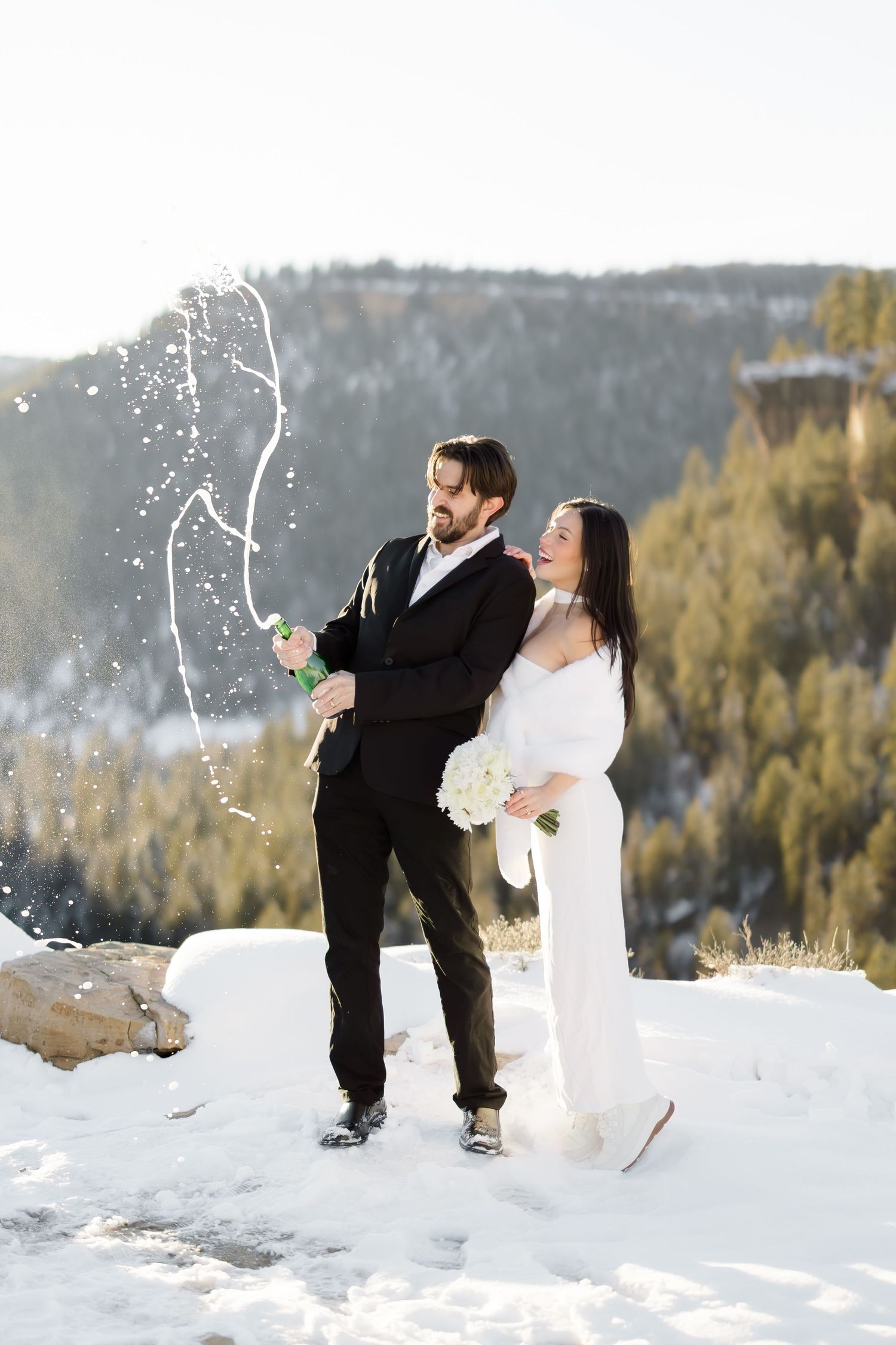 Joyful newlyweds celebrate with champagne spray on snowy mountain overlook in winter wedding.