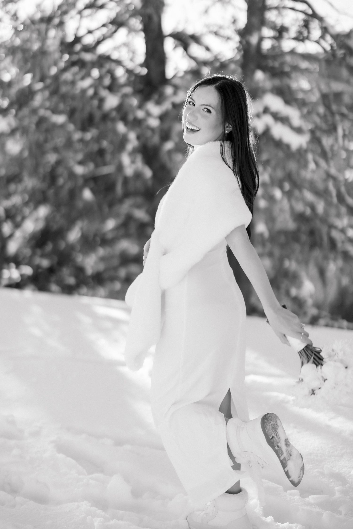 Black and white photo of smiling bride in white gown and fur stole walking through a snowy winter forest.