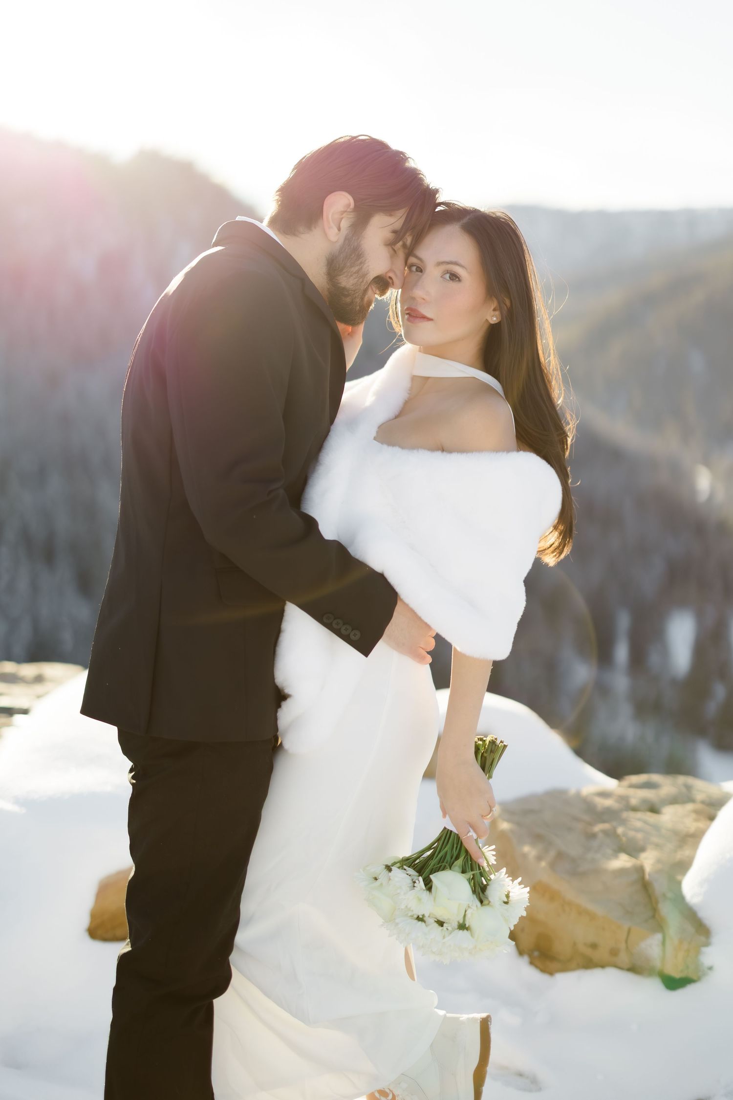 Bride and groom embrace in snowy mountain landscape, bride in white fur stole holding white bouquet.