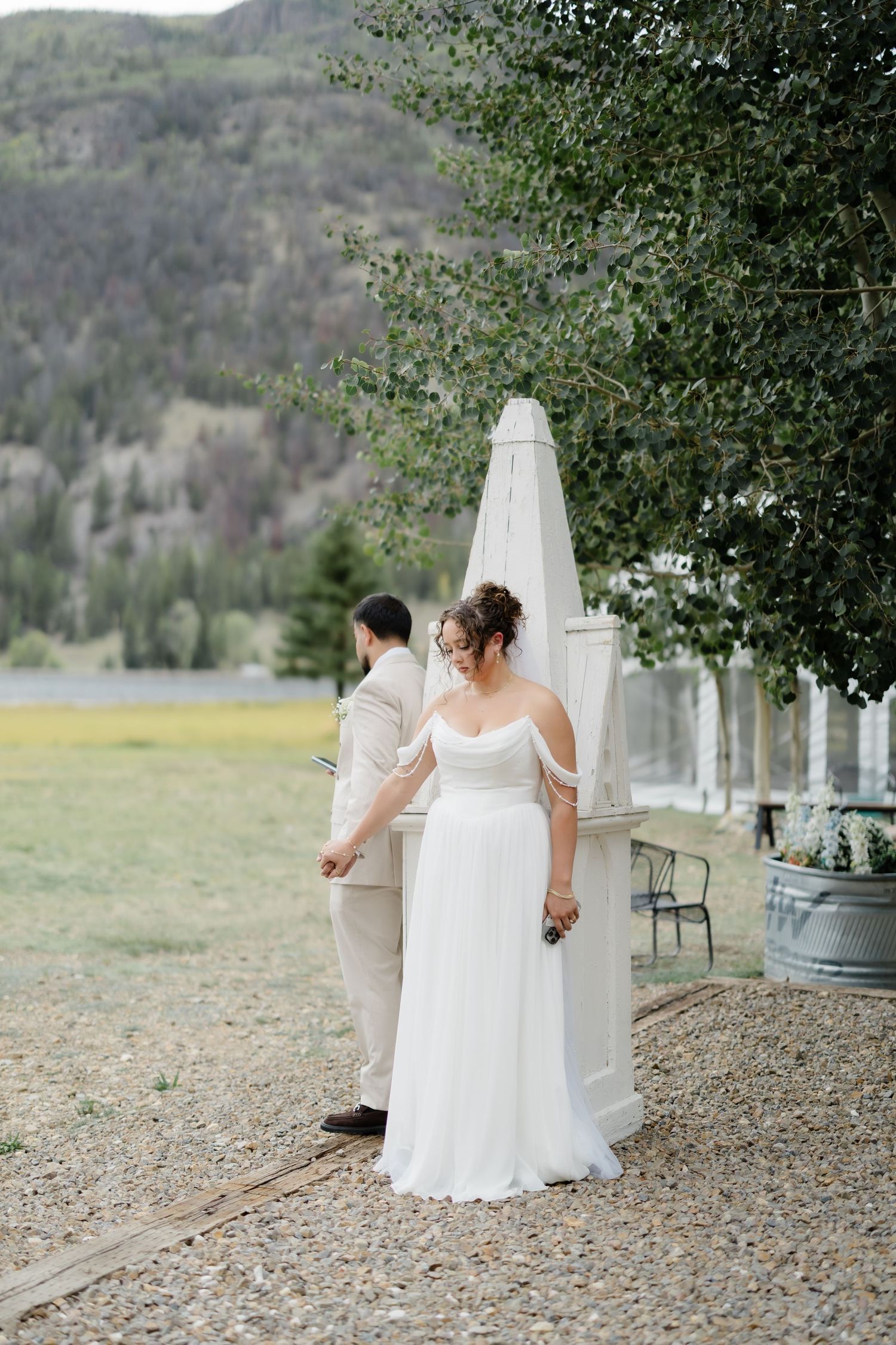 Bride in white off-shoulder gown and groom in tan suit share a first look at an outdoor mountain wedding venue.