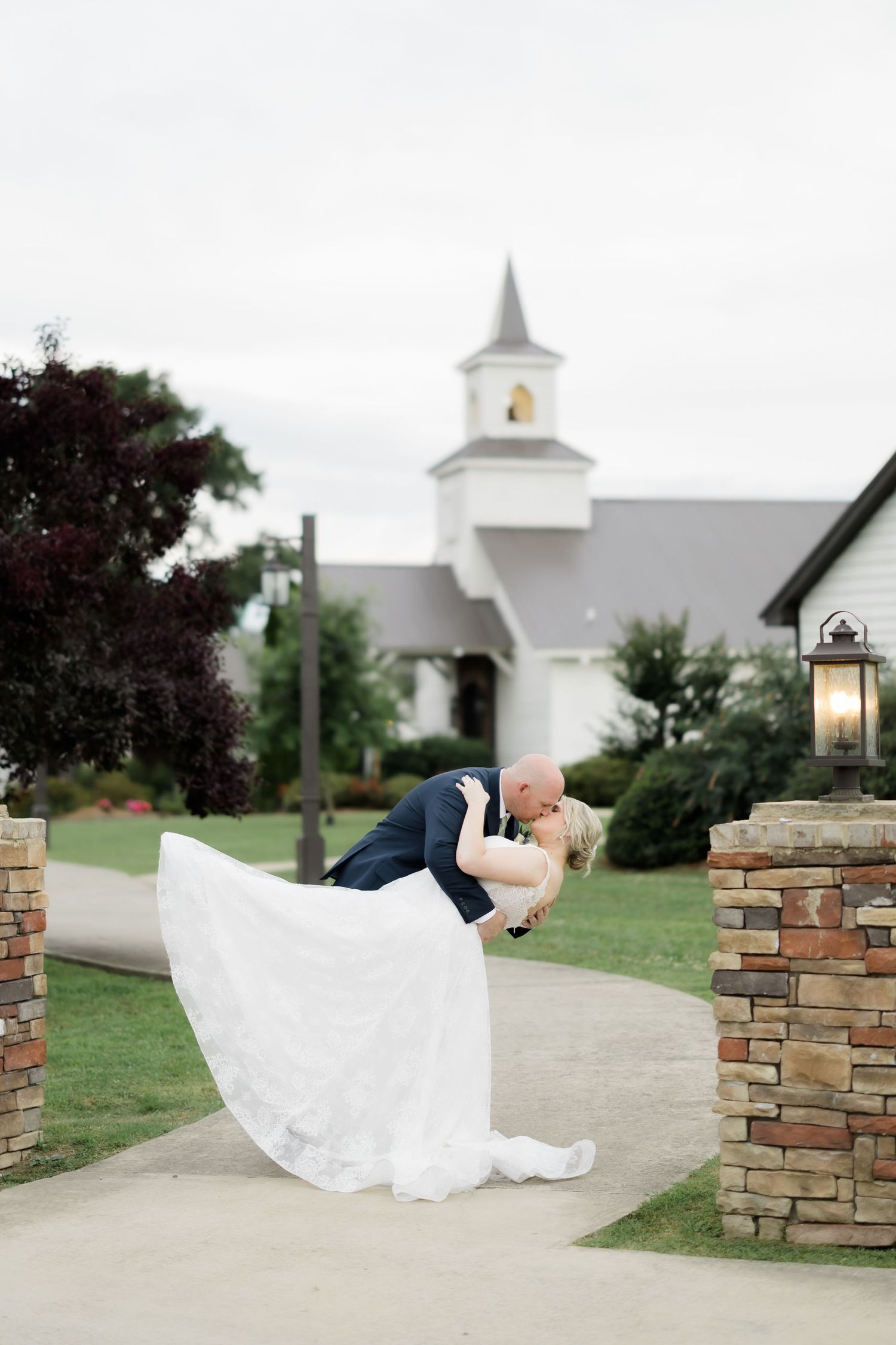 A groom dips his bride in a romantic kiss outside a white chapel, her flowing wedding gown sweeping gracefully.
