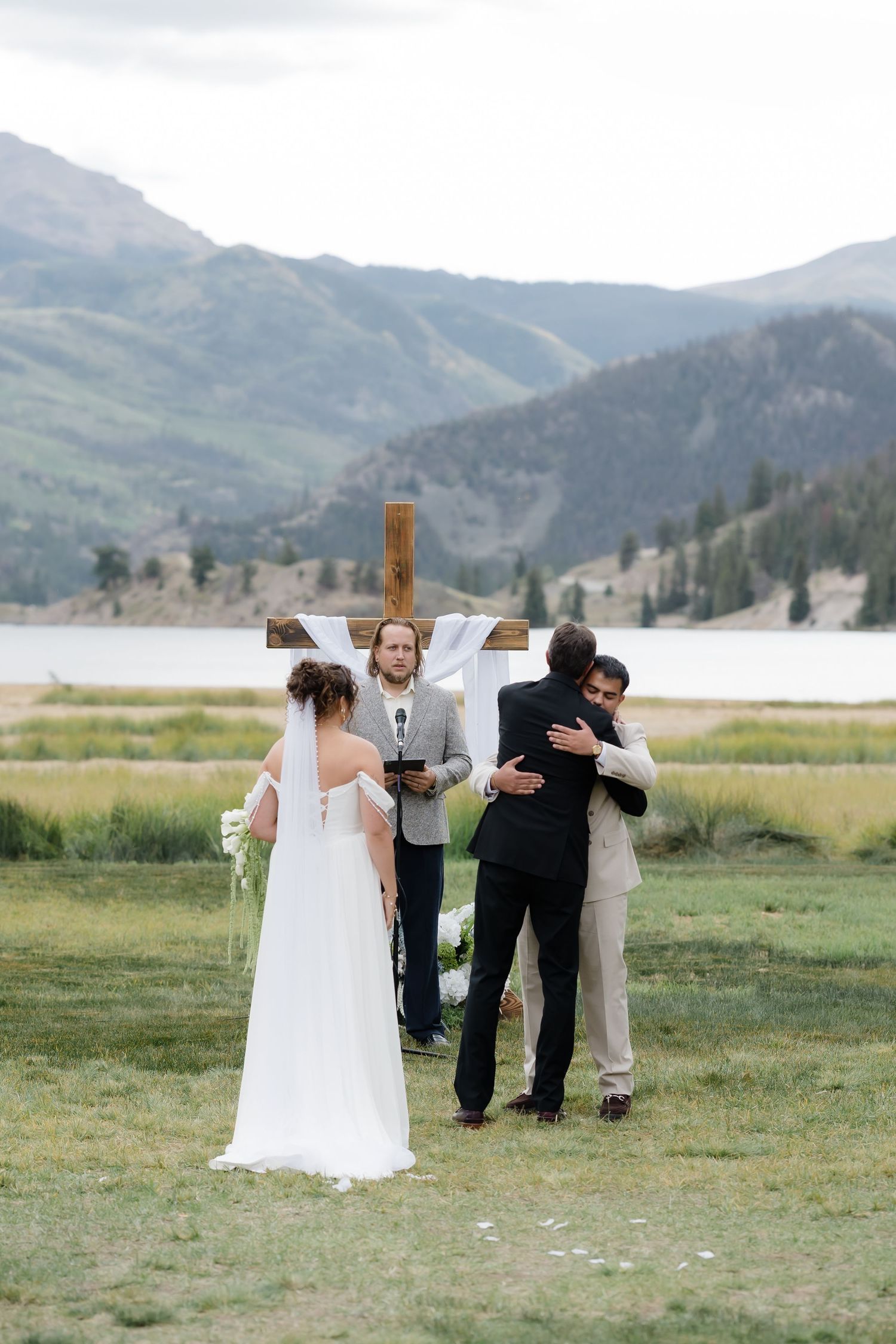 Outdoor mountain wedding ceremony with bride and groom hugging officiant before a wooden cross near a scenic lake.