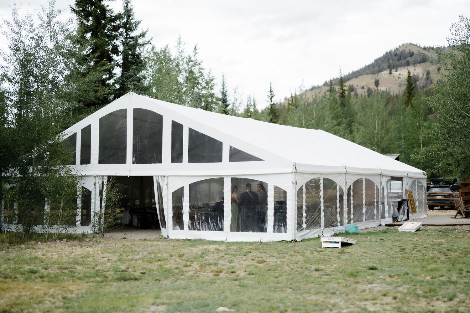 Large white event tent with clear walls set in a mountain meadow surrounded by pine trees and hills.