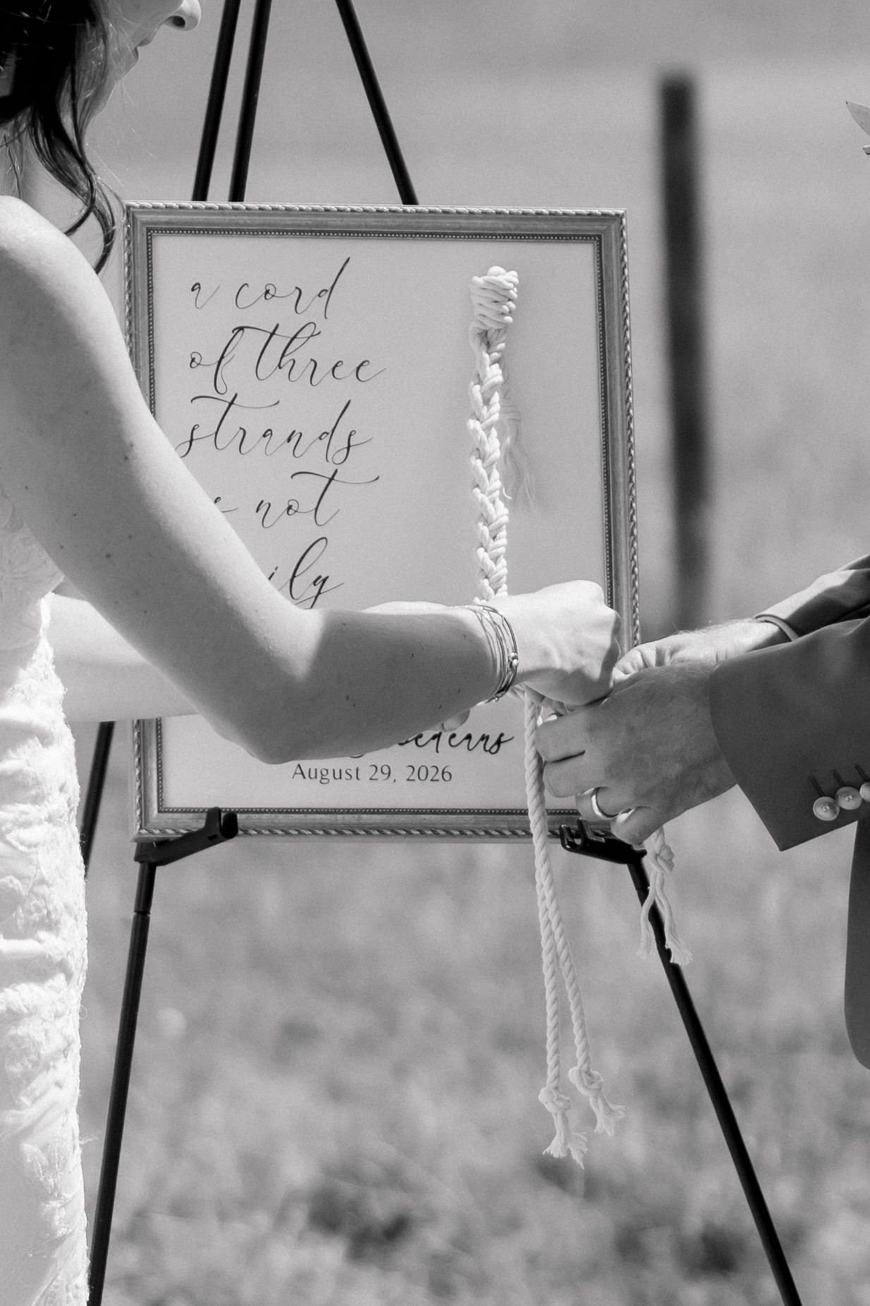 Black and white wedding photo of couple holding braided cord unity ceremony rope before framed calligraphy sign.