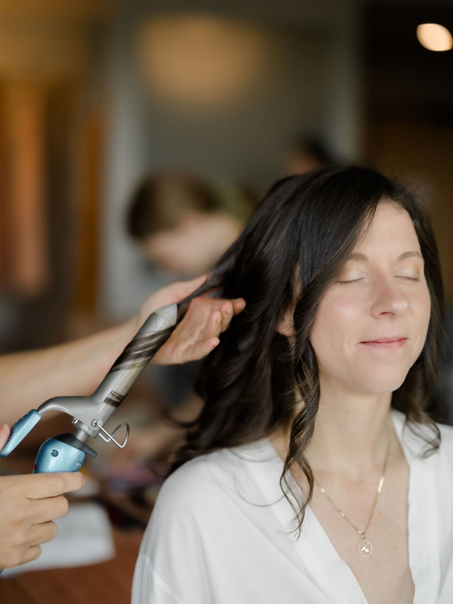 A woman with dark hair smiles peacefully while a stylist curls her hair with a curling iron.