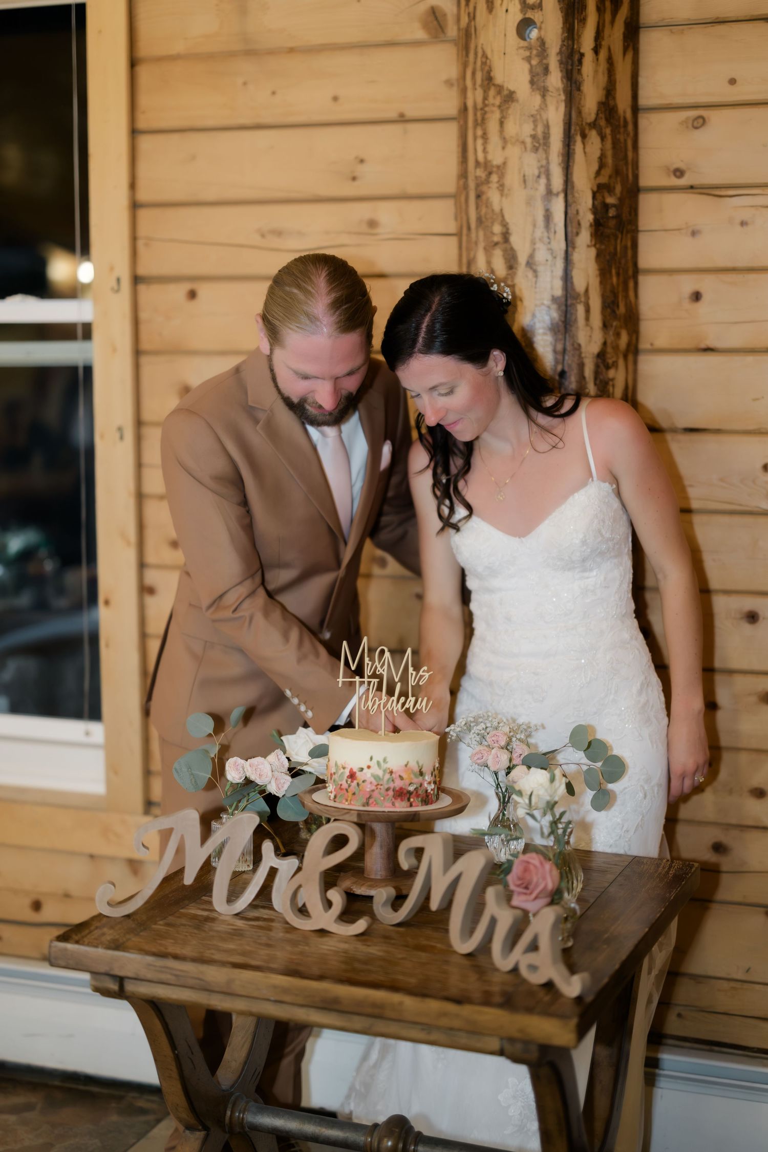 Bride and groom cutting wedding cake on rustic table with Mr & Mrs sign and floral decorations.