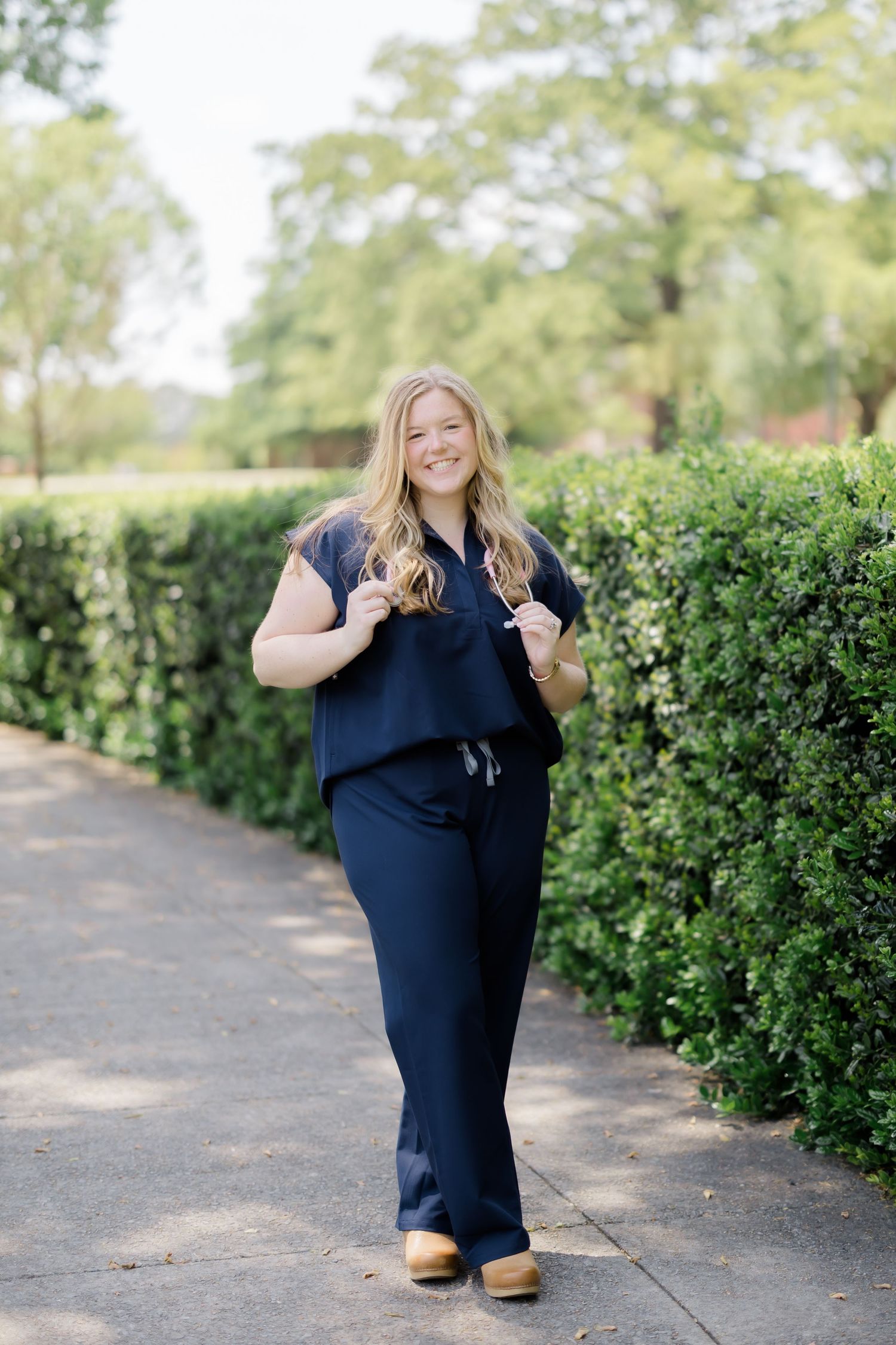 Woman in navy blue matching set and wooden clogs posing on a garden path with green hedges.