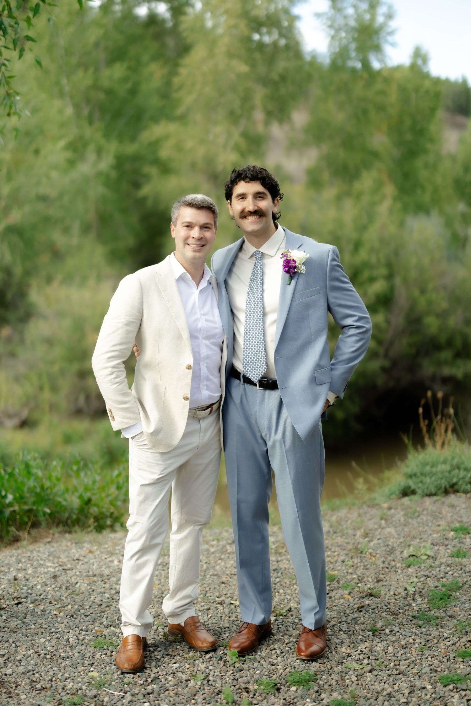 Two grooms in suits smile outdoors on gravel path with lush green trees behind them at their wedding.