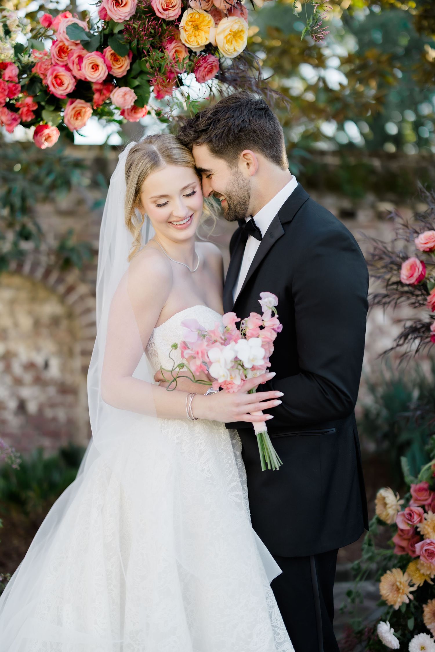 A romantic wedding scene with vibrant pink and yellow floral arrangements against a garden wall.